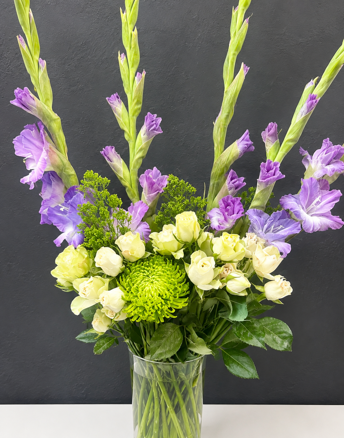 Custom floral arrangement featuring lavender gladiolus, white roses, green chrysanthemums, lavender flowers, and seasonal greenery.