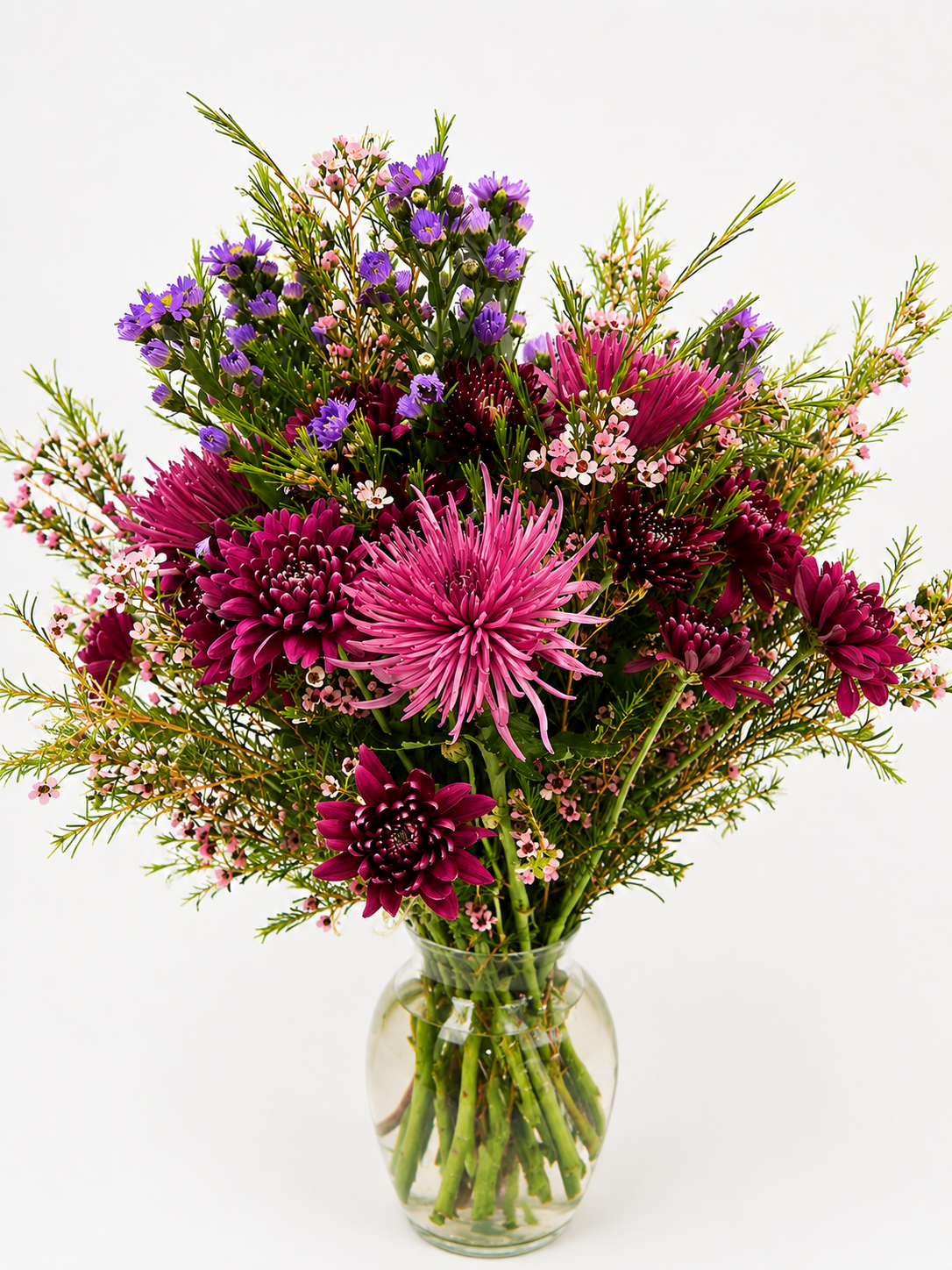 Lush floral arrangement featuring deep purple chrysanthemums, lavender flowers, pink accents, and delicate branching greenery.