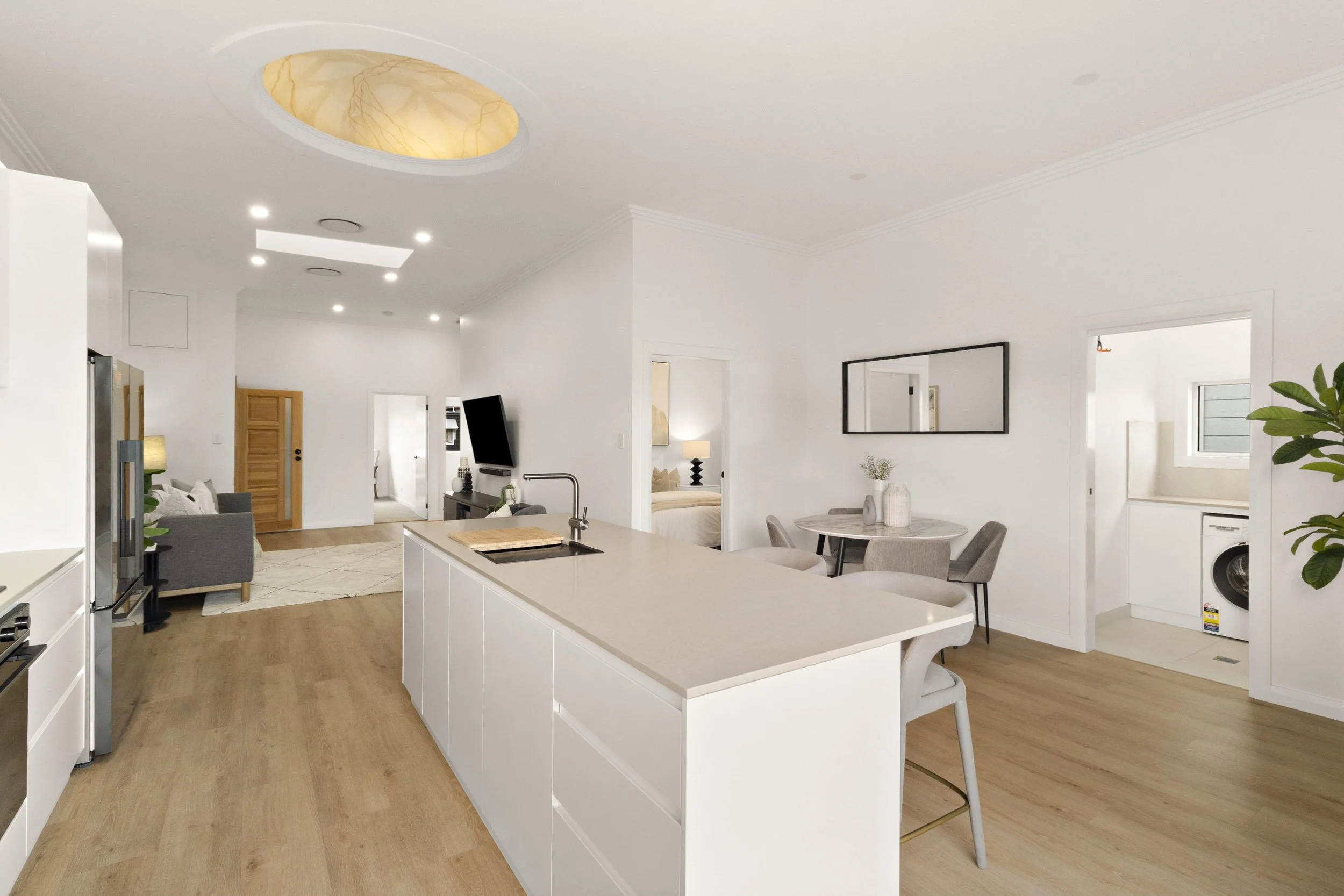 Open-concept kitchen and living area with white walls, wood flooring, and modern furniture. The kitchen has a large white island with a sink, and a round dining table with four chairs. The sitting area includes a television, a gray armchair, and a small sofa.