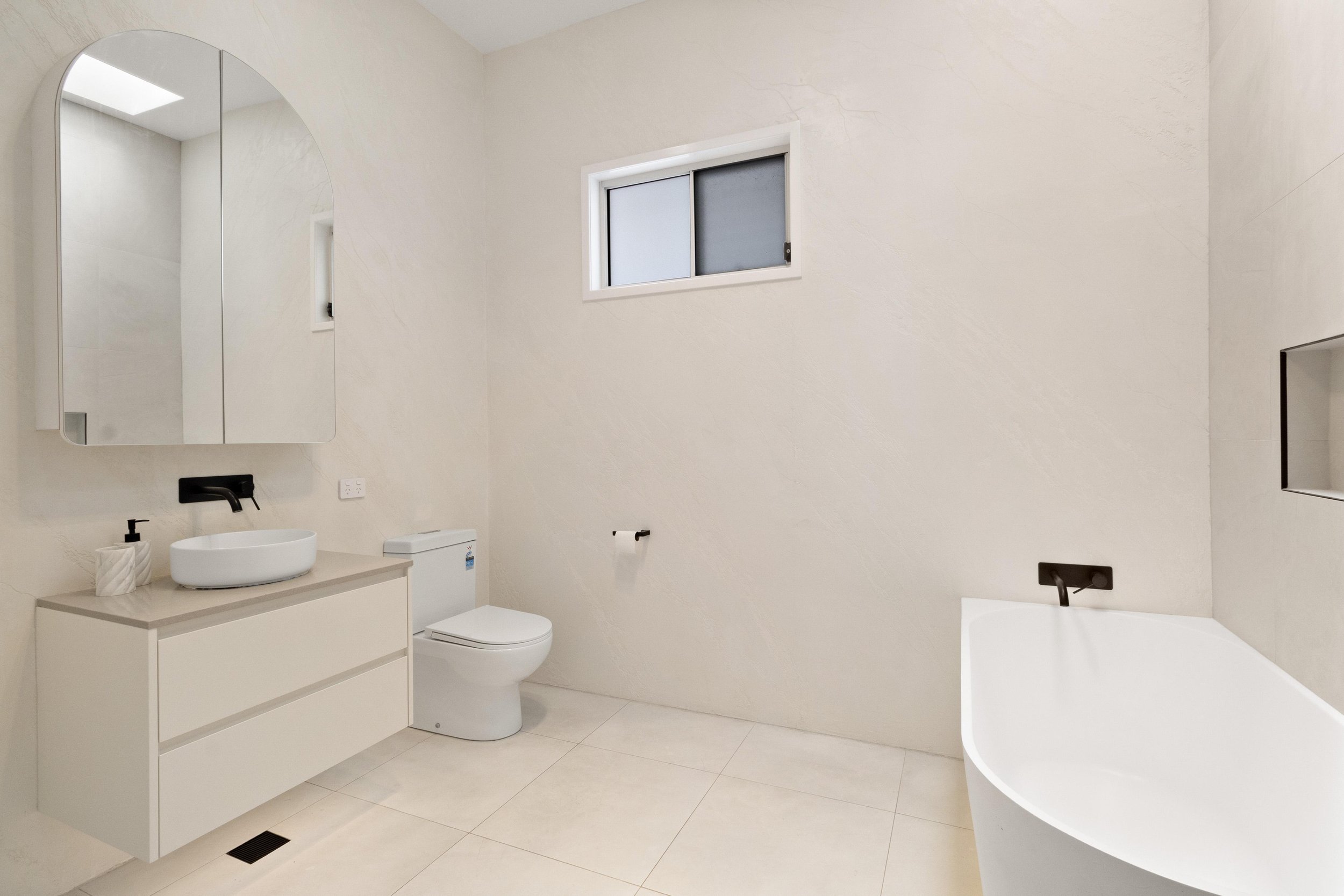 Modern minimalist bathroom with a white floating vanity, above counter sink, wall mirror, toilet, small window, and bathtub, all in neutral tones.