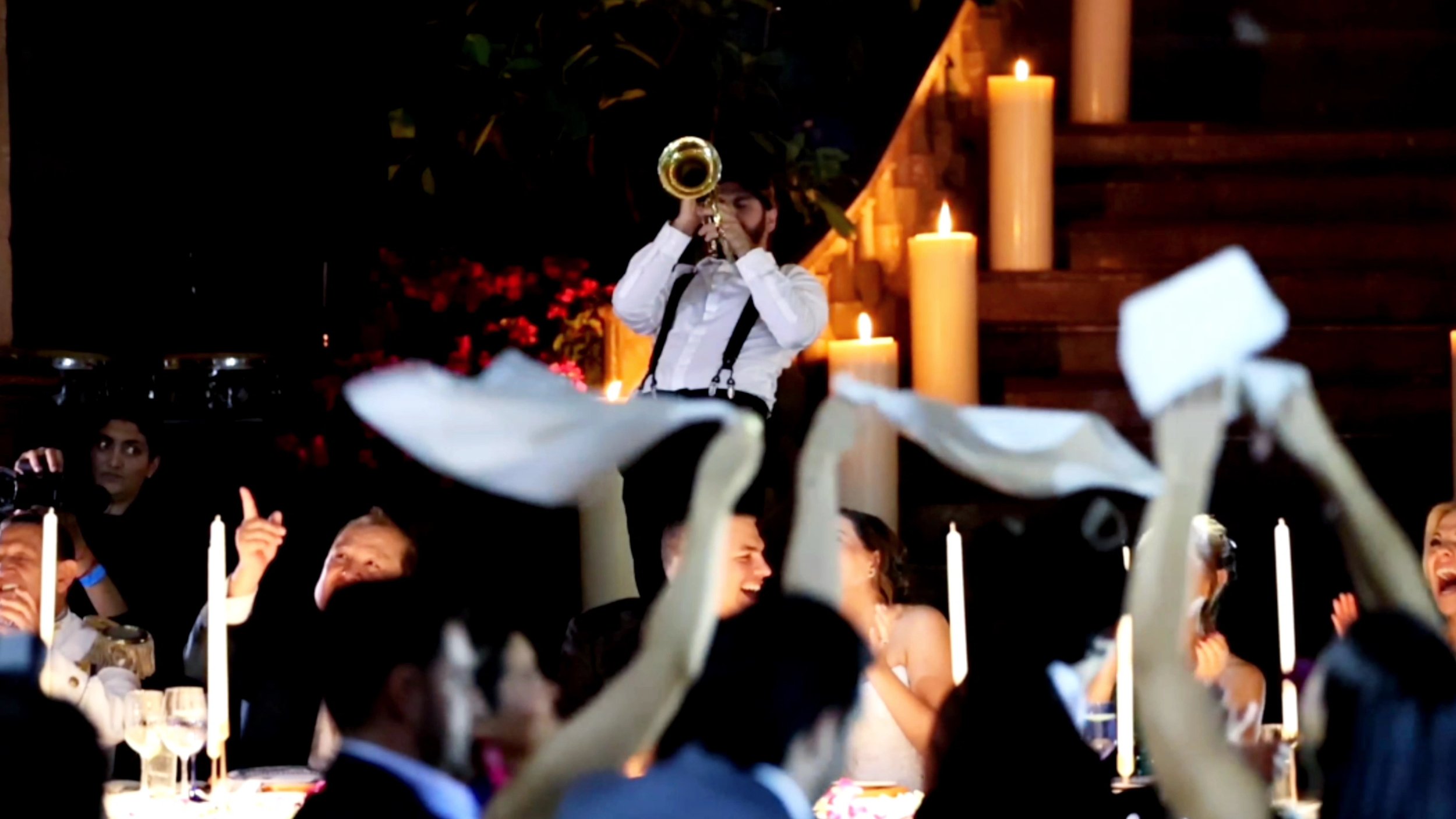 Hombre tocando trompeta en una celebración o boda con invitados bailando y decoraciones con velas y flores