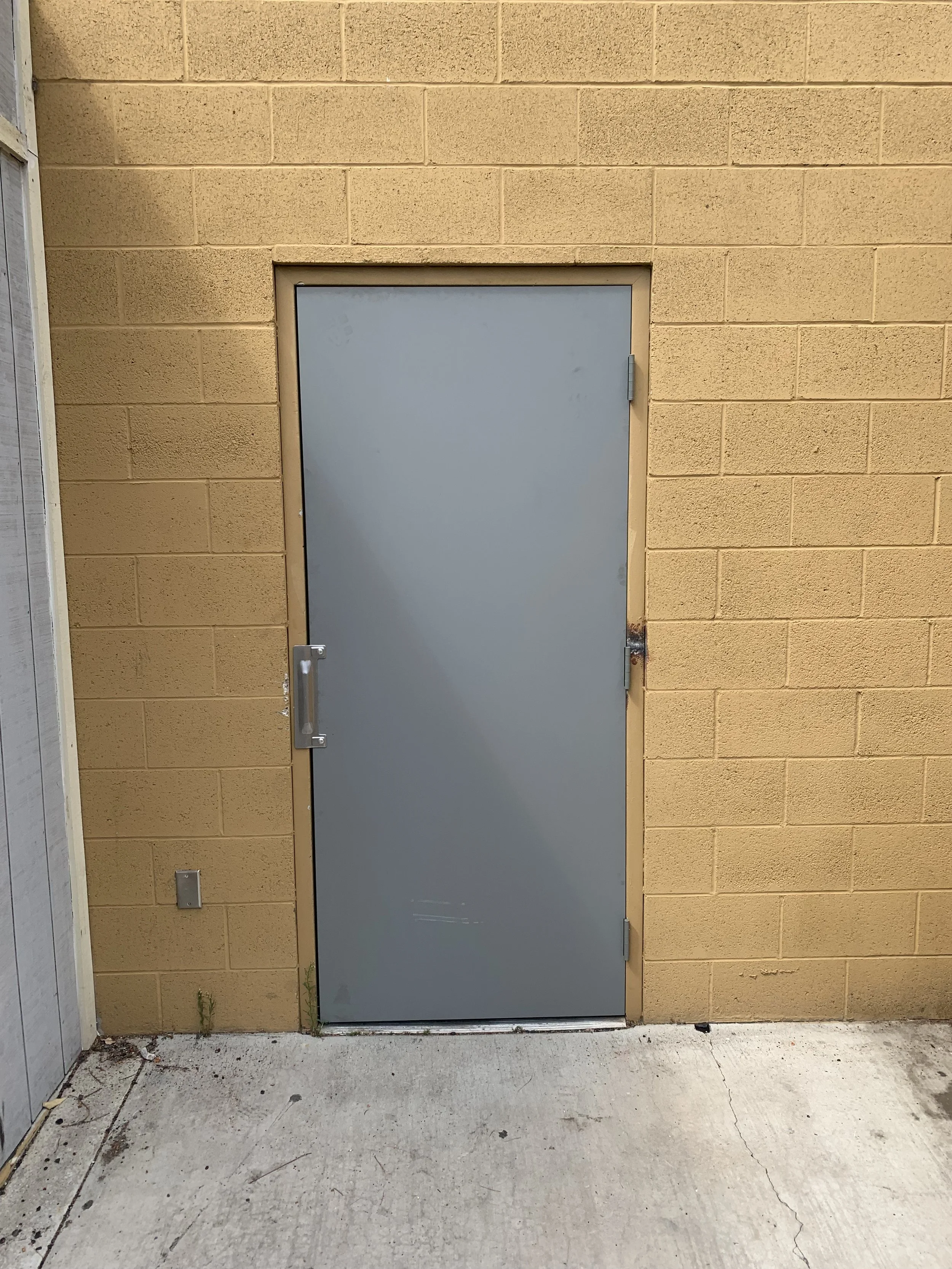 Gray metal door with a handle, set into a yellow brick wall exterior, on concrete ground.
