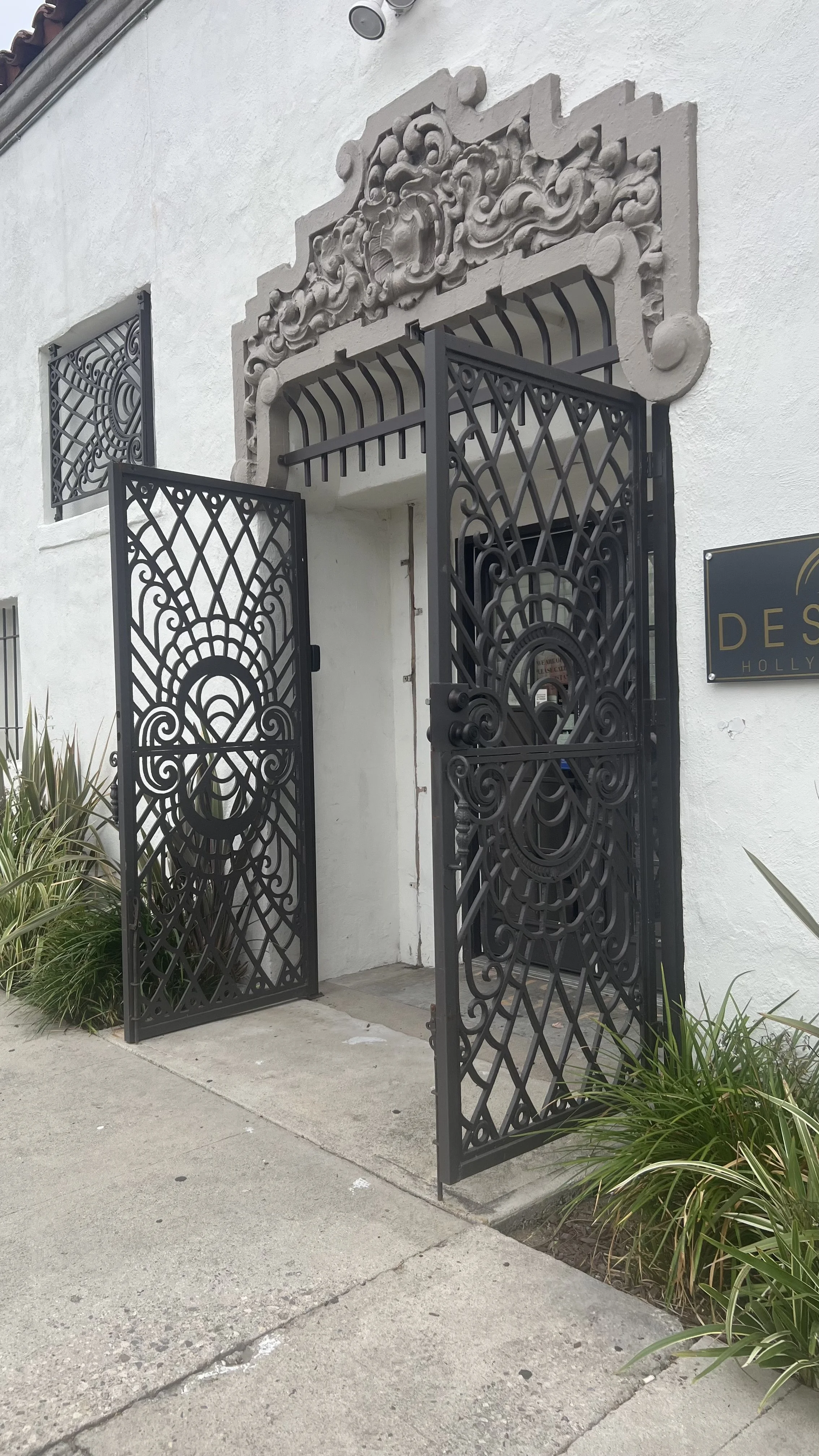 Black ornate wrought iron gate open in front of a white building with decorative stone archway above the entrance and a small window with iron grill on the side. Some plants are visible near the gate.