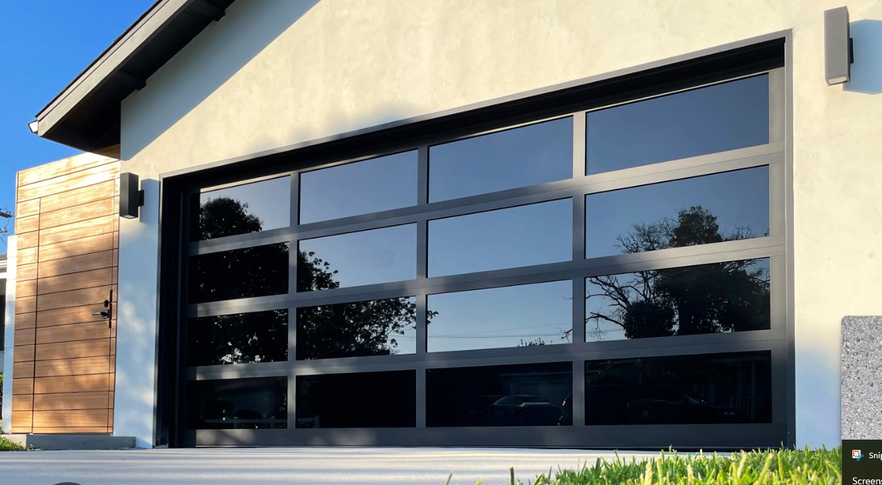 Modern garage door on a light-colored house with a small wooden accent wall and a concrete walkway, reflecting trees and a blue sky.