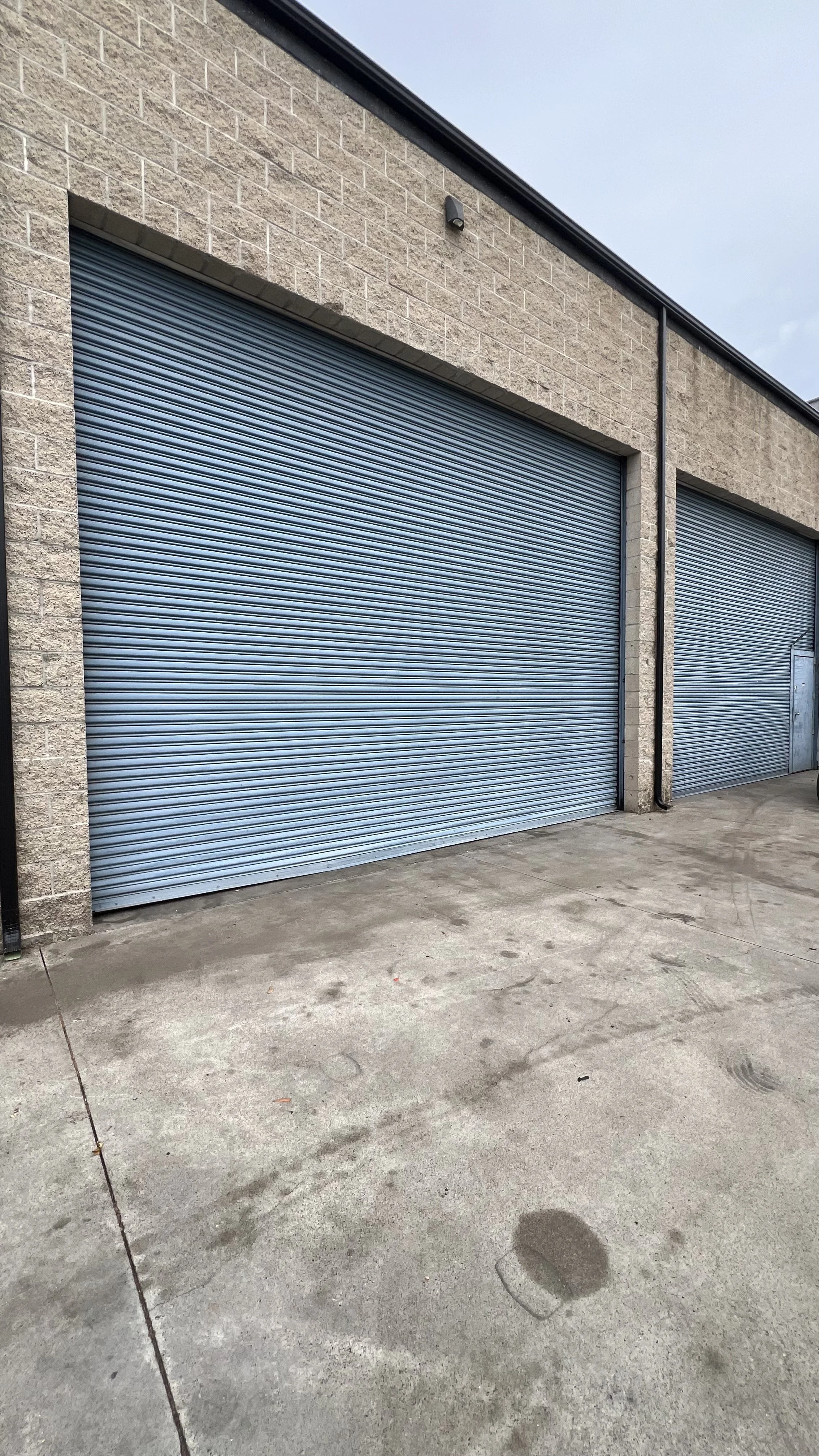 Two closed blue metal roll-up garage doors in a brick building with concrete pavement in front.