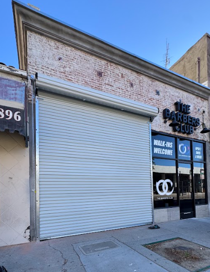 storefront with a closed metal roll-up door, signage for The Barbers Club, and an entrance with glass doors and windows displaying the logo and signs for walk-ins welcome