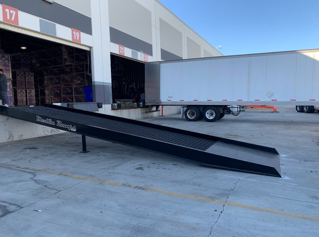 Loading dock with a black loading ramp labeled 'Medlin Ramp & Co.' extending from a warehouse door to the ground, with a white semi-trailer truck parked nearby.