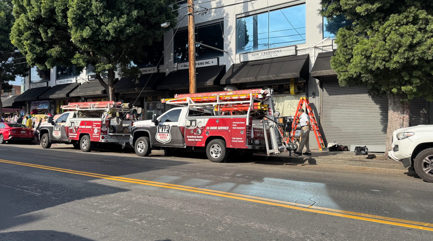 Two service trucks parked on a city street in front of a storefront, with one worker setting up safety cones and equipment, and several bags on the sidewalk. The storefront has black awnings and signs for fabric and foam store and other businesses.
