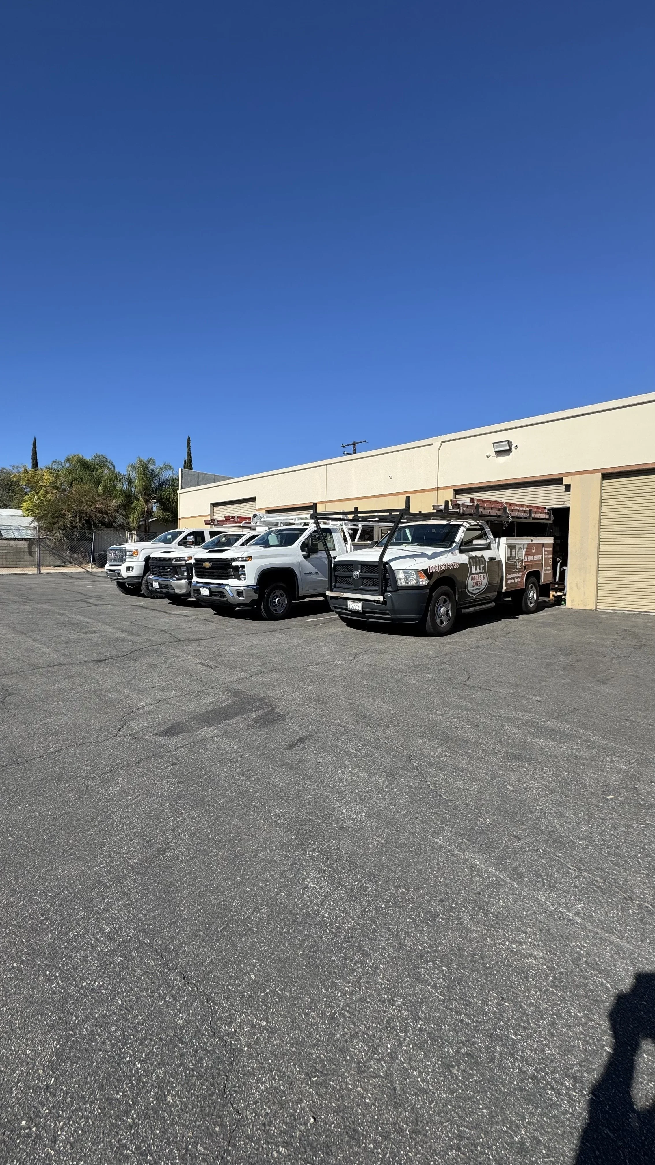 A parking lot with four white service trucks parked in front of a beige building with roll-up doors. Clear blue sky and some greenery are visible in the background.