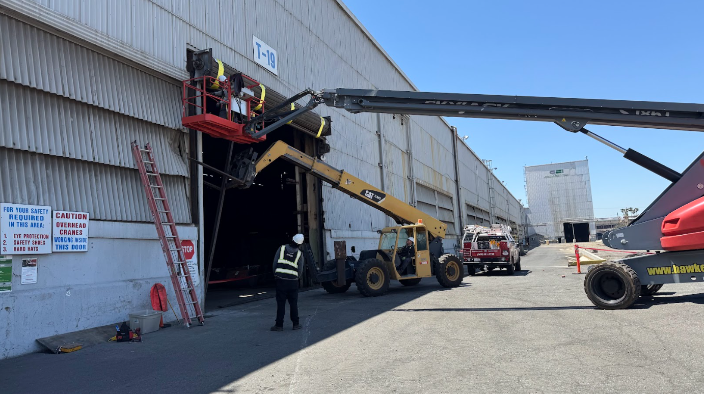 Workers and machinery inspecting the entrance of a large industrial building with a damaged or open garage door, safety signs posted on the wall, and a clear blue sky overhead.