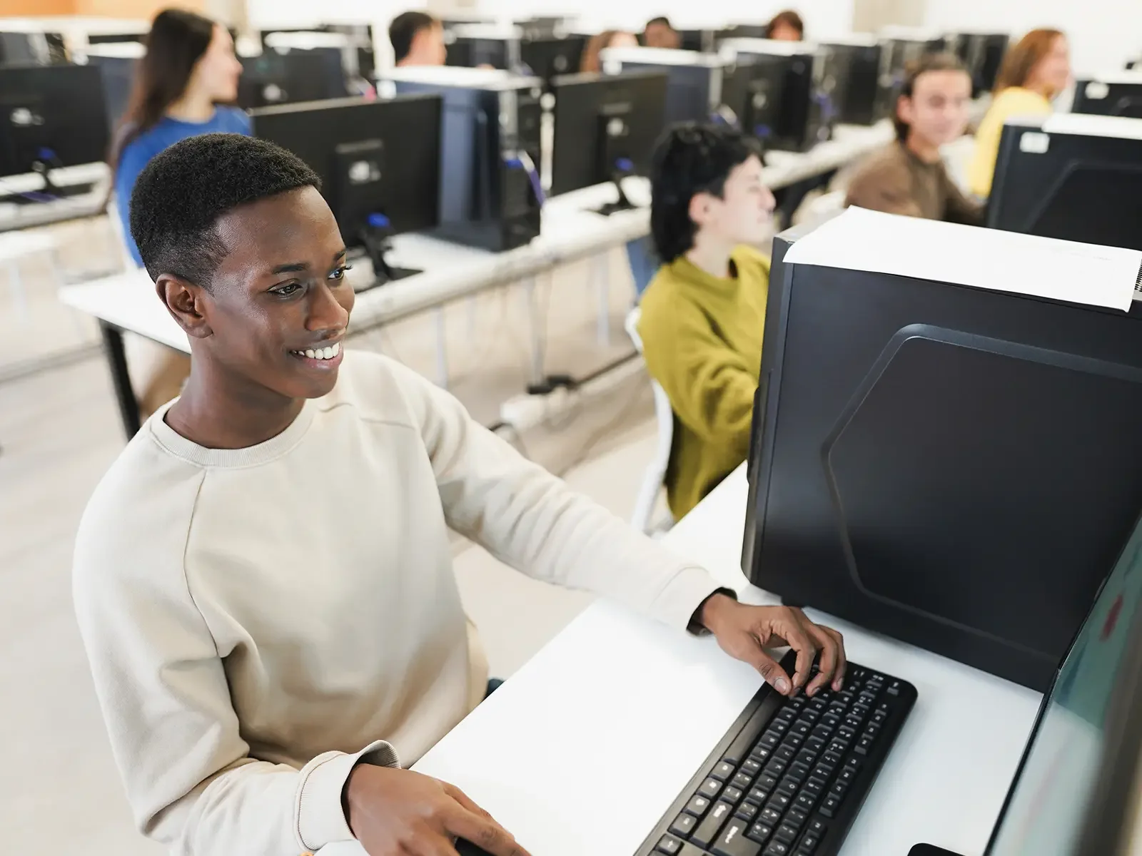 High school student working at a desktop computer in a public school computer lab with classmates in the background