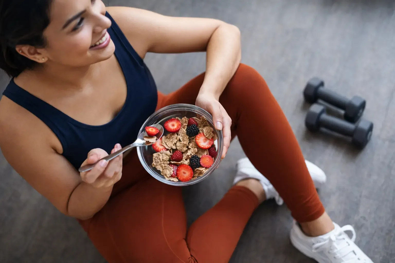 Woman sitting on the floor at a gym, holding a bowl of cereal with strawberries, blackberries, and cereal flakes, while smiling and looking to the side. Dumbbells are on the floor nearby.