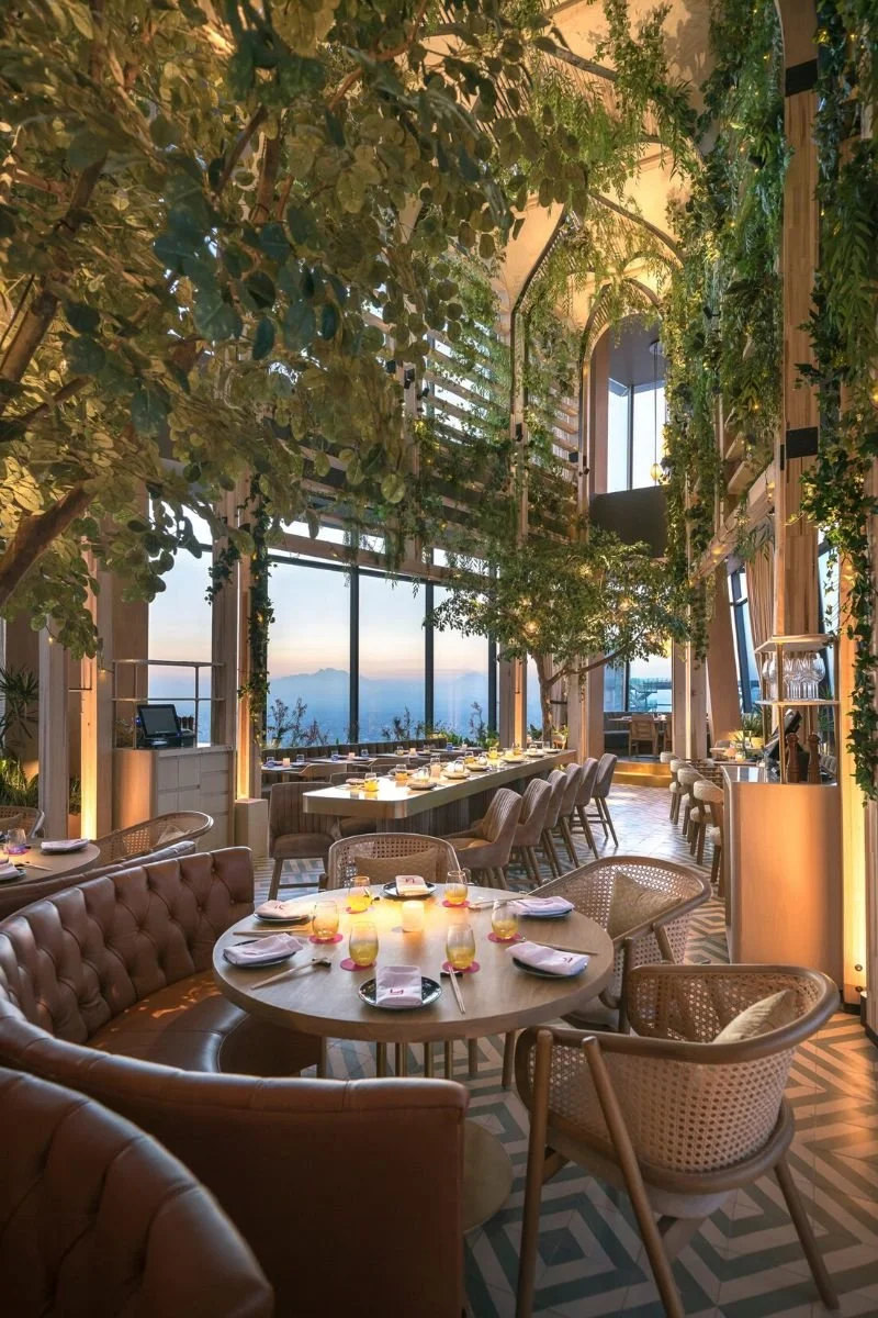 Elegant restaurant interior with large windows, trees, and greenery, set for dining with tables, chairs, and place settings.