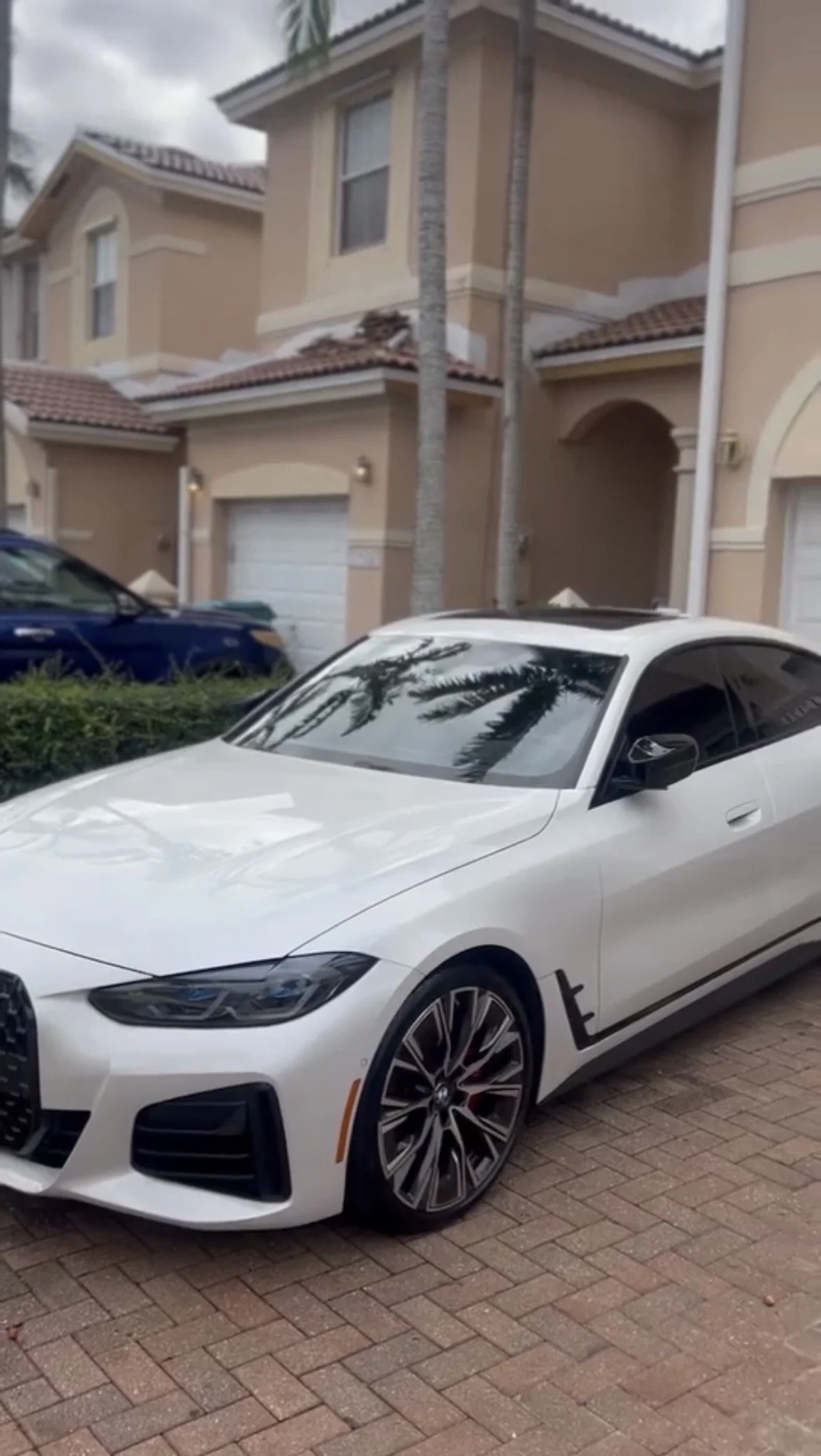 A white sports car parked on a brick driveway in front of a beige townhouse with palm trees reflected in the windshield.
