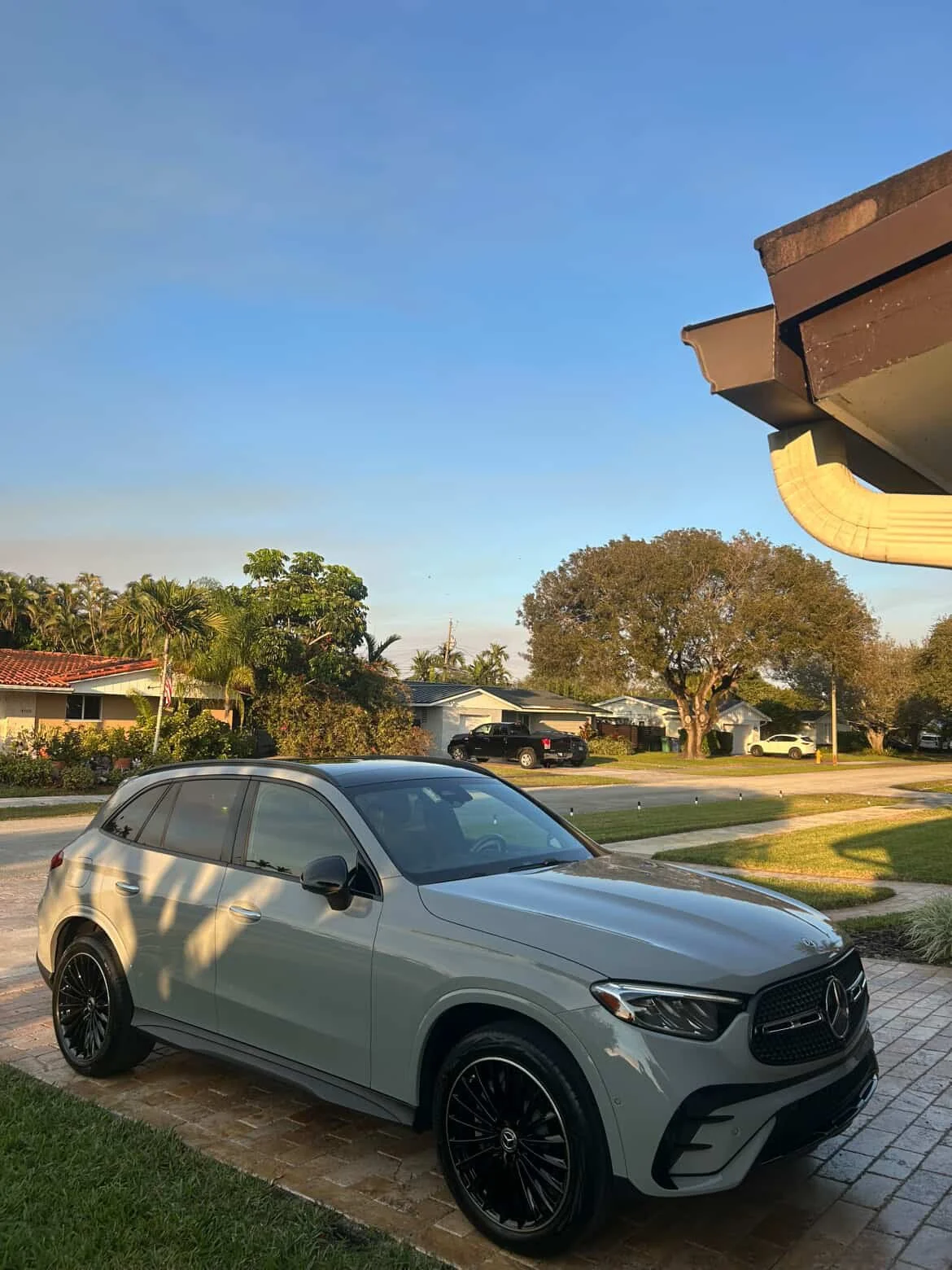 A silver Mercedes-Benz SUV parked on a brick driveway in front of a suburban house during sunset with palm trees and other houses in the background.
