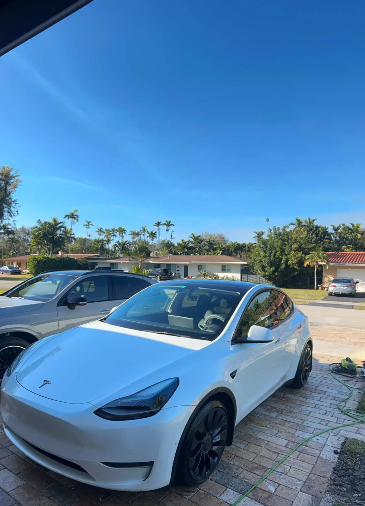 A white Tesla Model Y parked on a brick driveway with a black roof and black wheels, reflecting nearby houses and trees on its surface, under a clear blue sky.