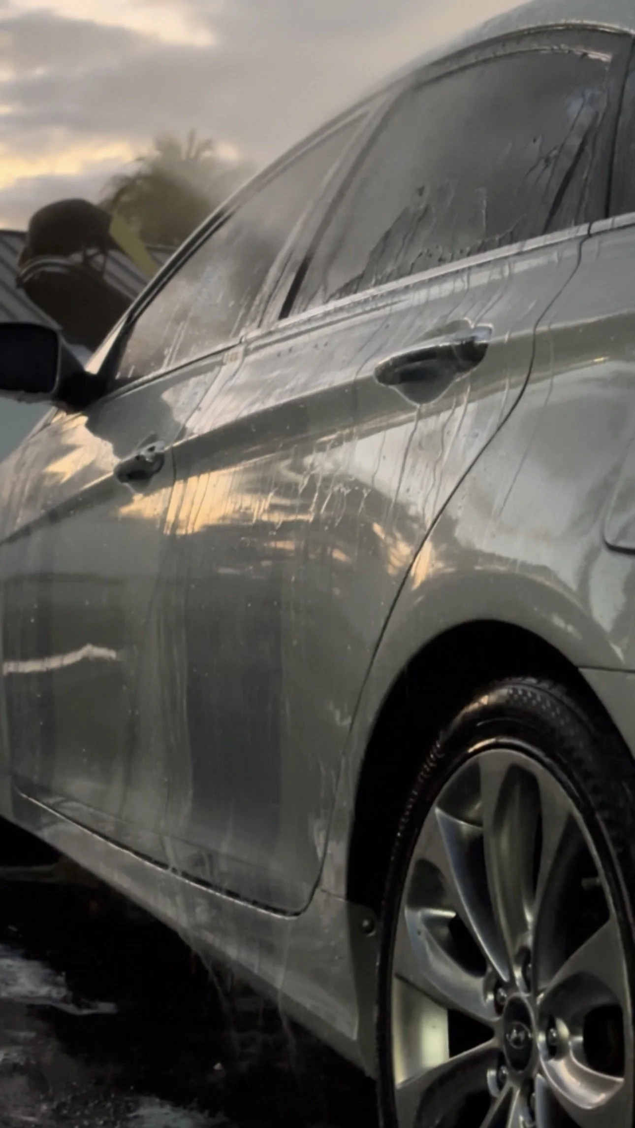 Side view of a silver car with water and dirt on its surface, damaged door, and a wet tire, under a cloudy sky.