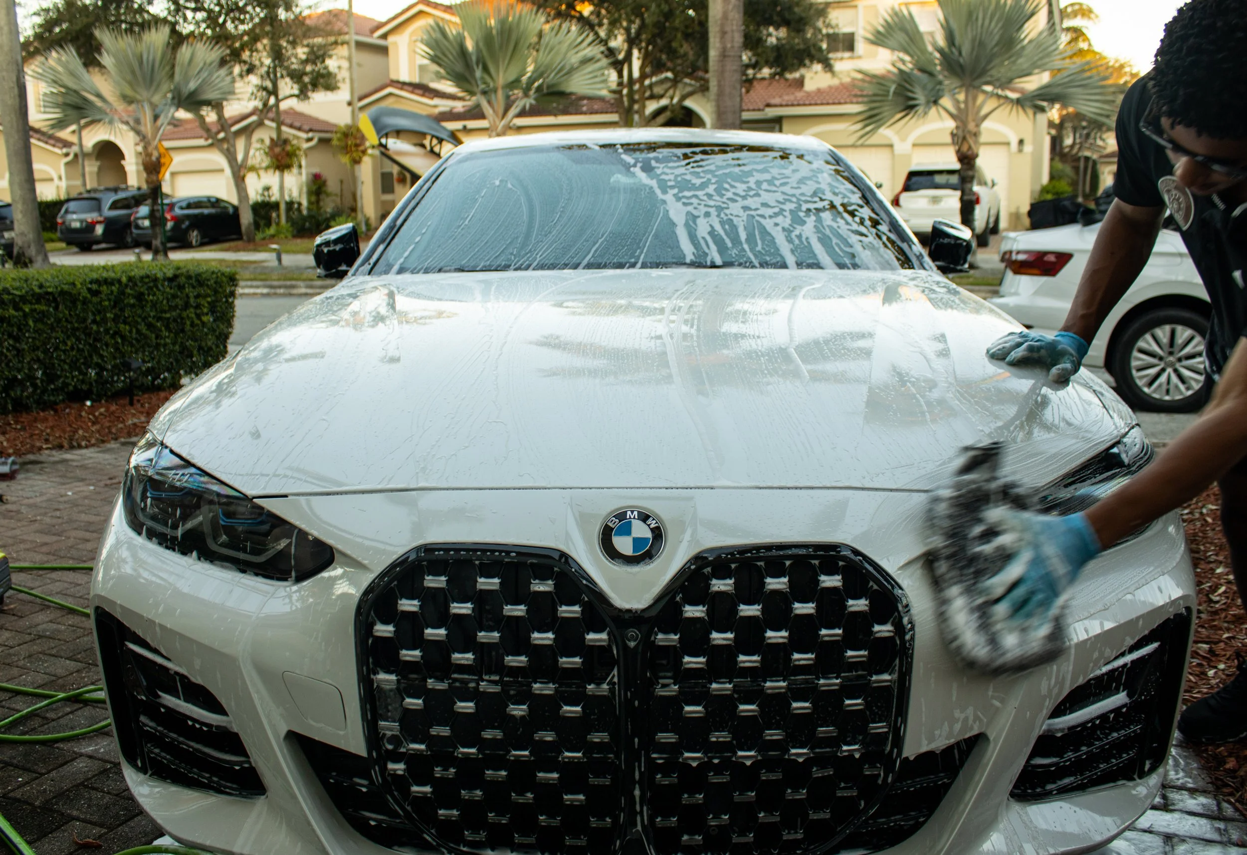 A person washing a white BMW car with soap and a sponge, outdoors in a residential area with palm trees and houses in the background.