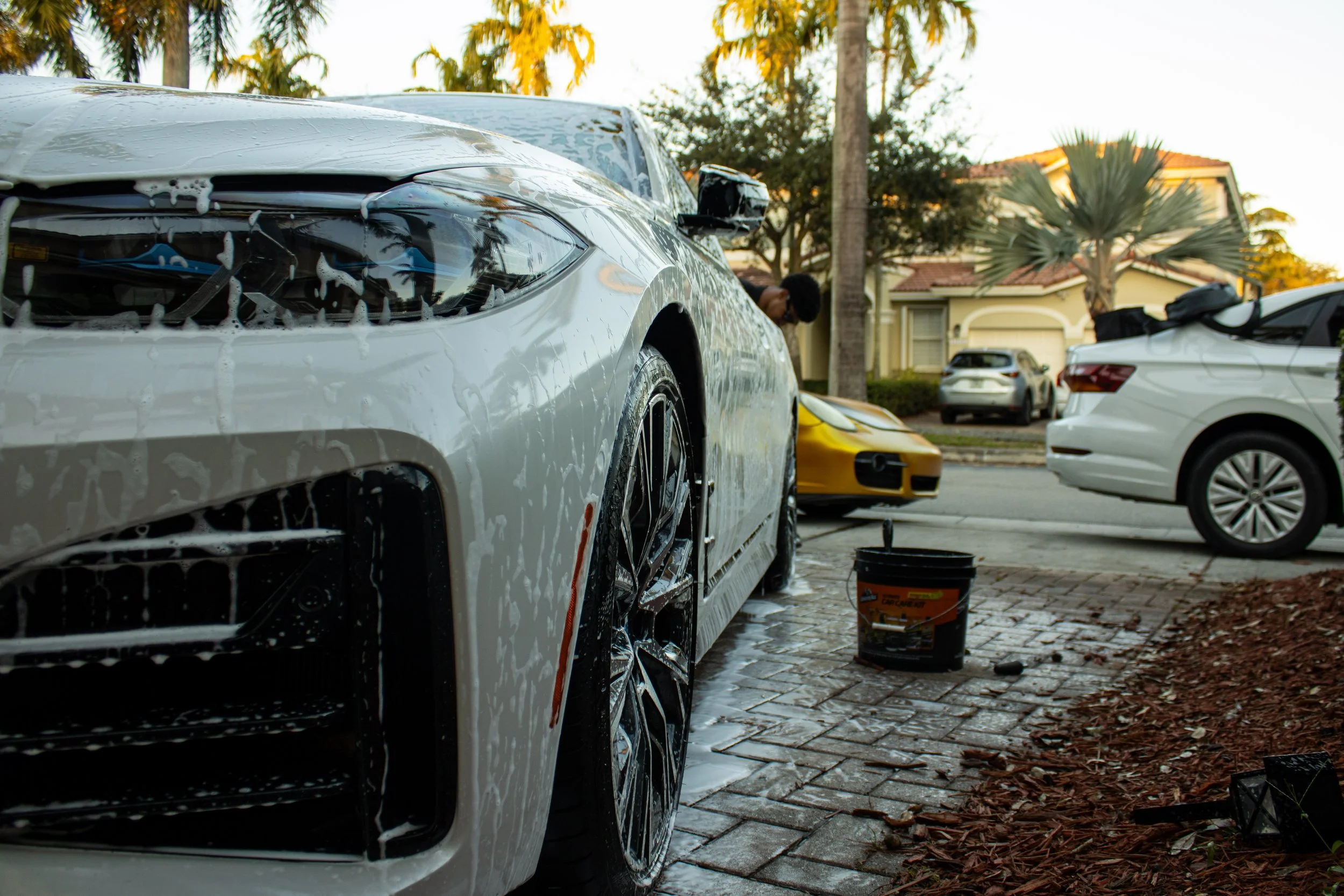 A white car being washed with soap suds, on a driveway in a suburb, with other cars parked, including a yellow sports car and a white sedan, and a person leaning over the yellow car.