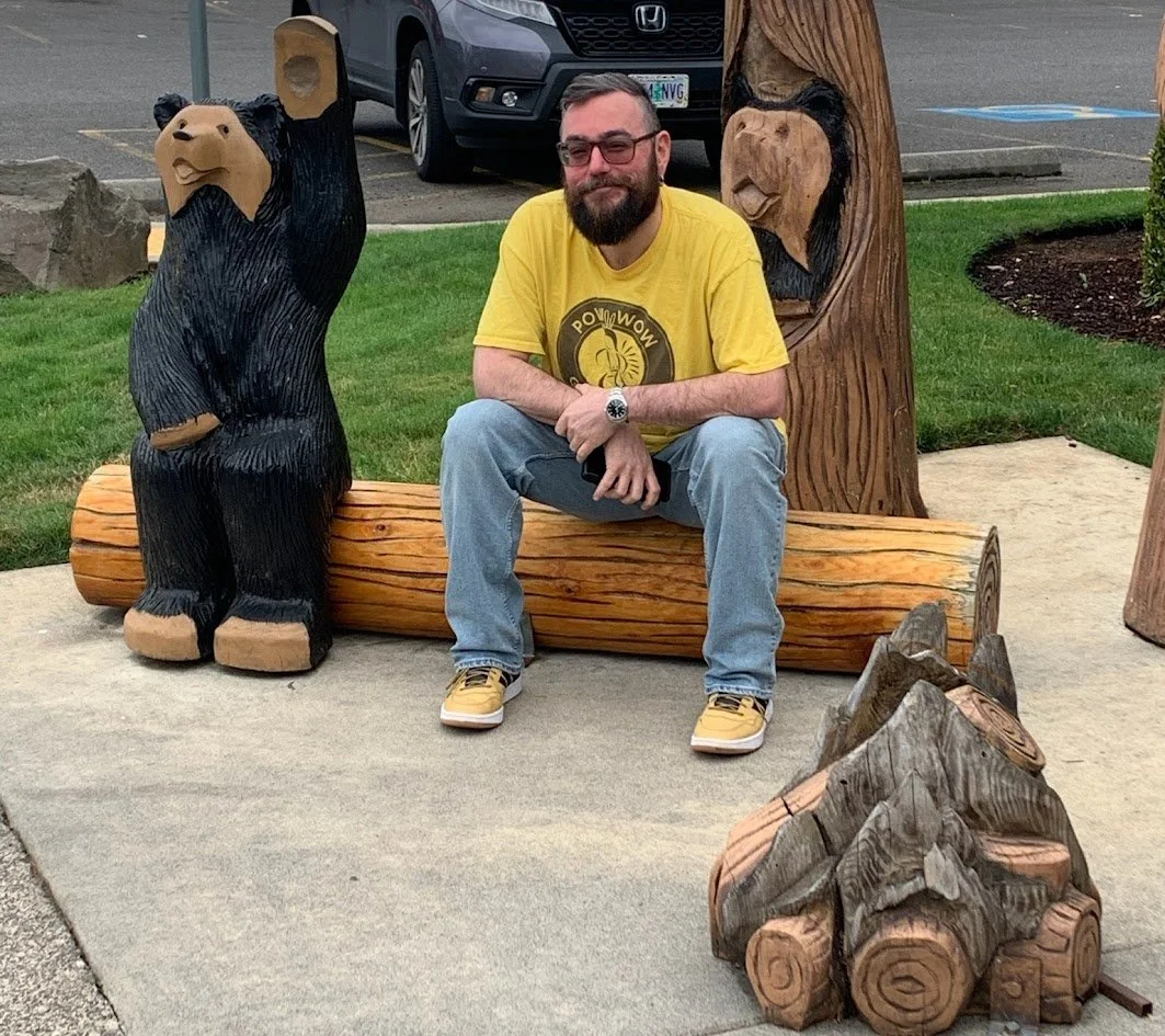 A man with a beard and glasses sitting on a wooden log bench, smiling. He is wearing a yellow t-shirt with a graphic and light blue jeans. He is surrounded by wooden sculptures of bears, including one to his left and one behind him. There are cars and a parking lot in the background.
