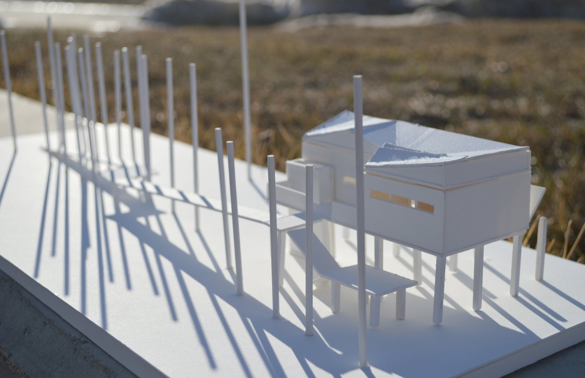 A detailed architectural model of a modern building with a flat roof, surrounded by white picket fences, placed on a white surface outdoors with grass and a road in the background.
