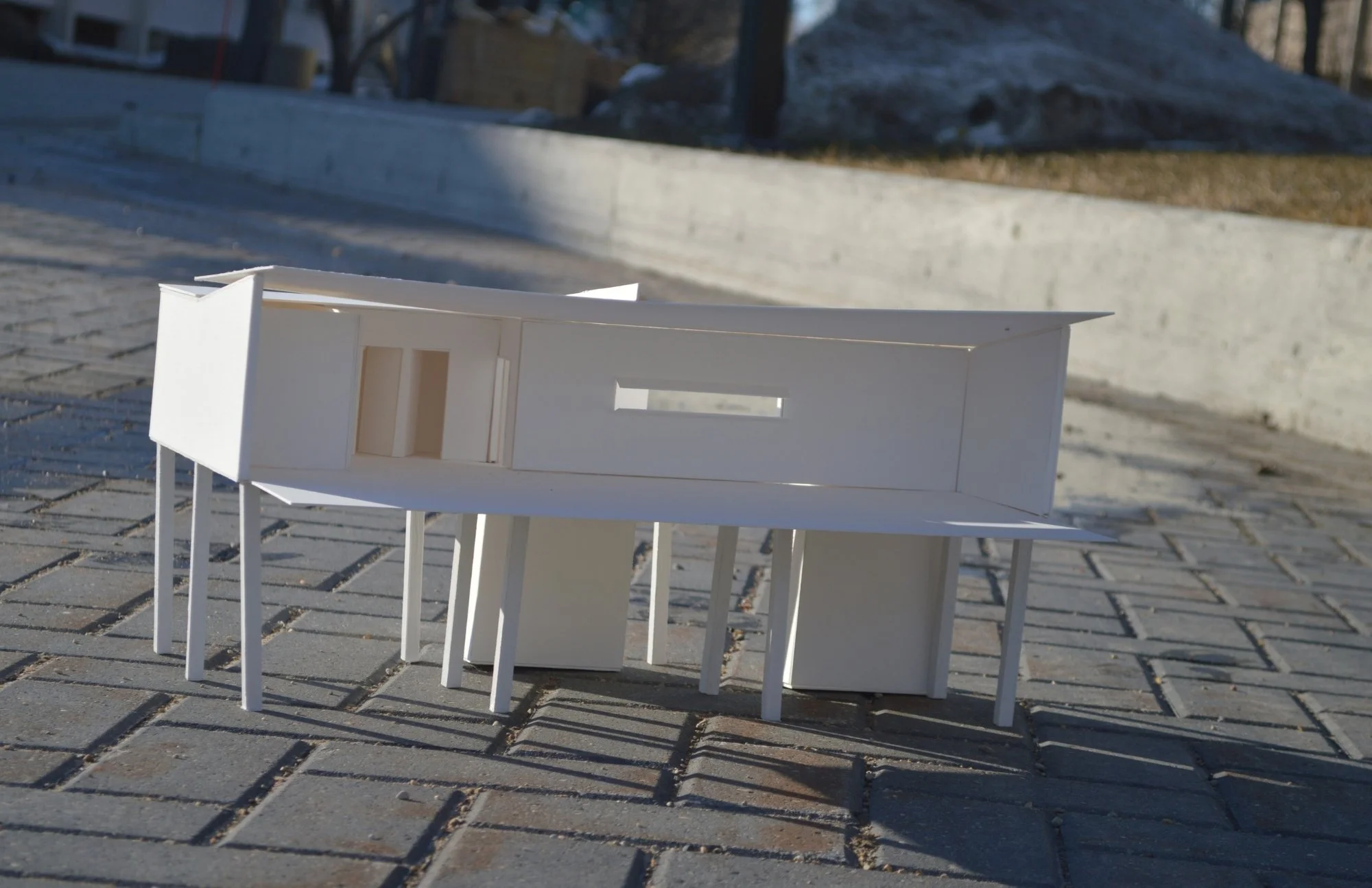 A small architectural model of a modern house placed outdoors on a paved surface with gravel and snow in the background.