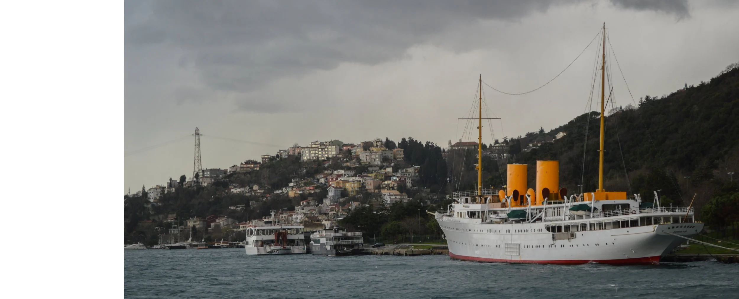 A large white historic ship with yellow funnels and masts docked in a harbor with a hillside cityscape in the background under cloudy skies.