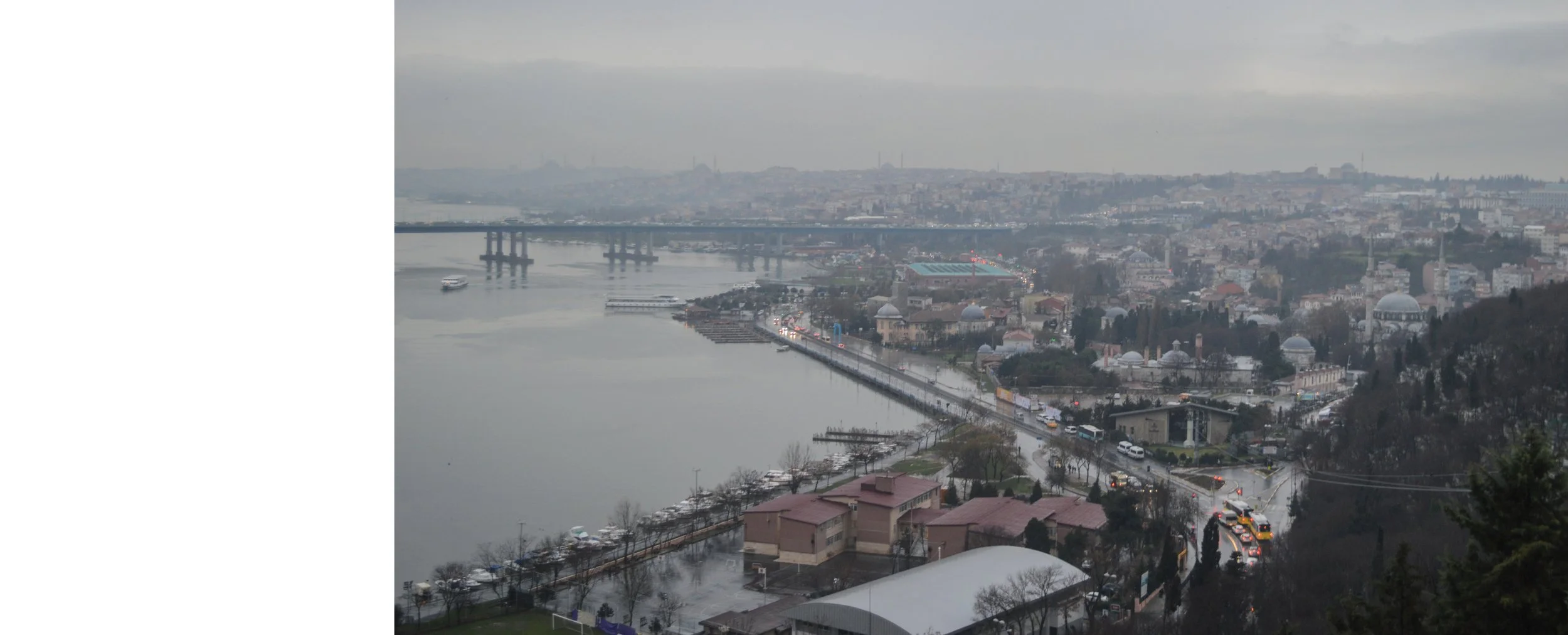 Overcast cityscape with a river, bridge, and buildings, viewed from a hill.