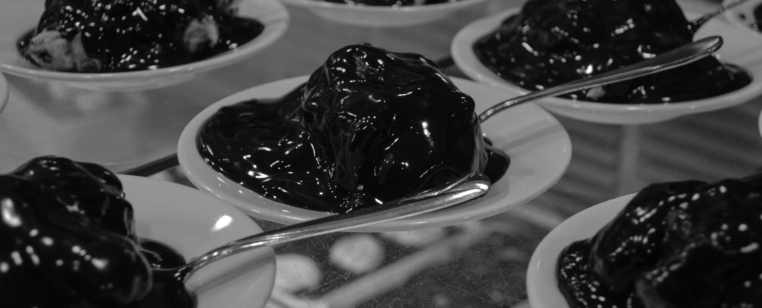 Small white dish filled with a glossy, dark jelly-like substance with a spoon inside. Multiple similar dishes with the same dark substance are visible in the background.