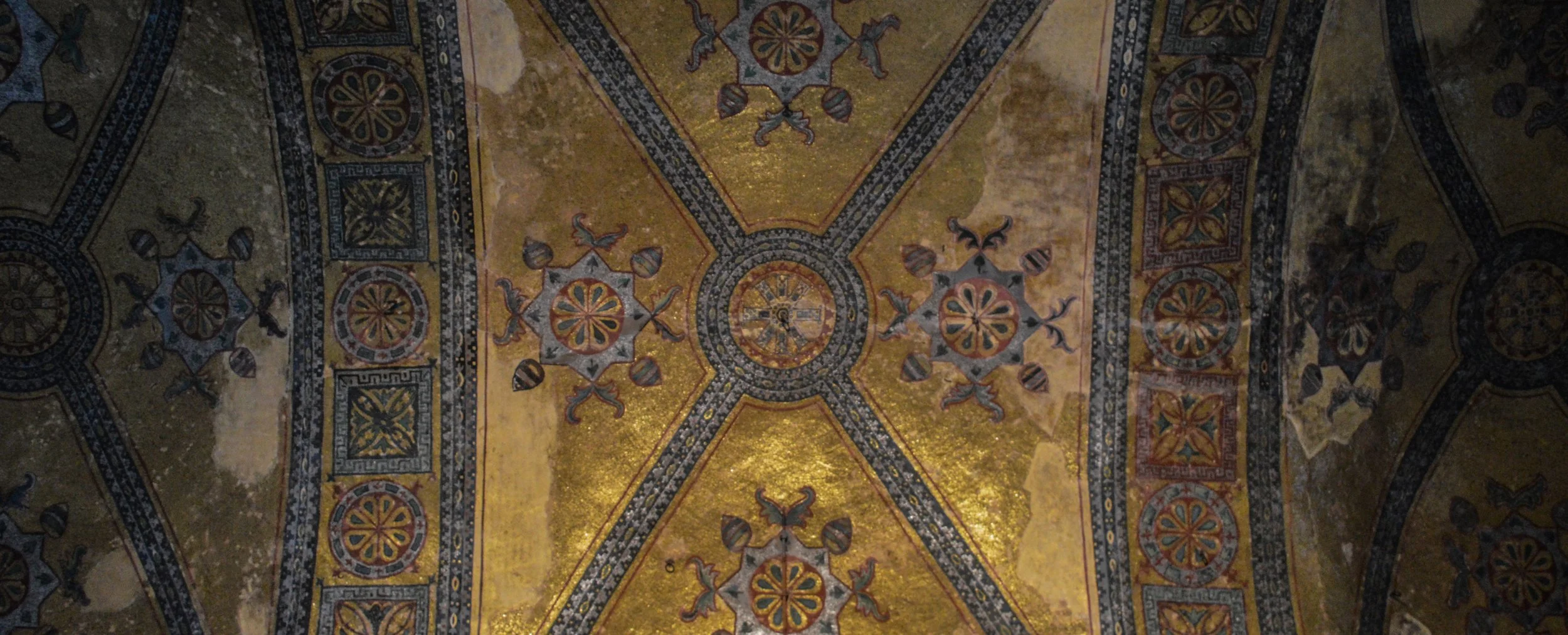 Ancient mosaic ceiling with intricate geometric and floral patterns in gold, blue, and red colors.