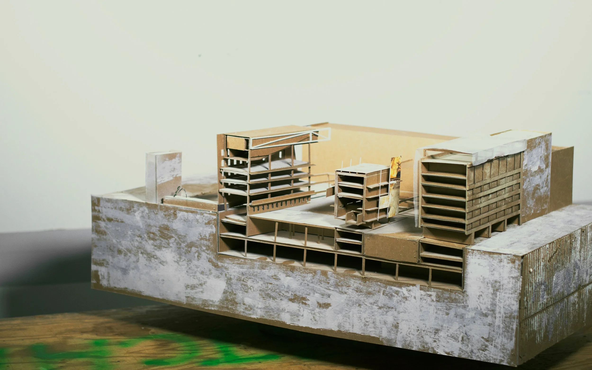 Architectural model of a multi-story building with various sections, made of cardboard and other materials, on a textured base.