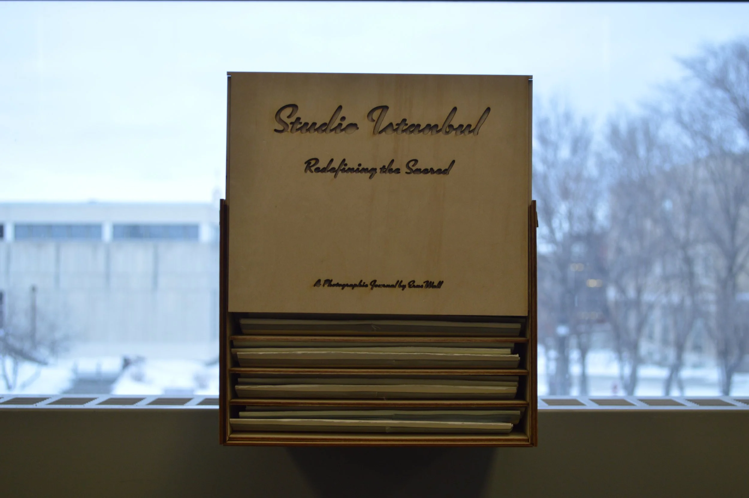 Book display stand with a sign reading "Studio Istanbul" and "Redefining the Sacred" against a backdrop of a window showing a snowy outdoor scenery with trees and buildings.