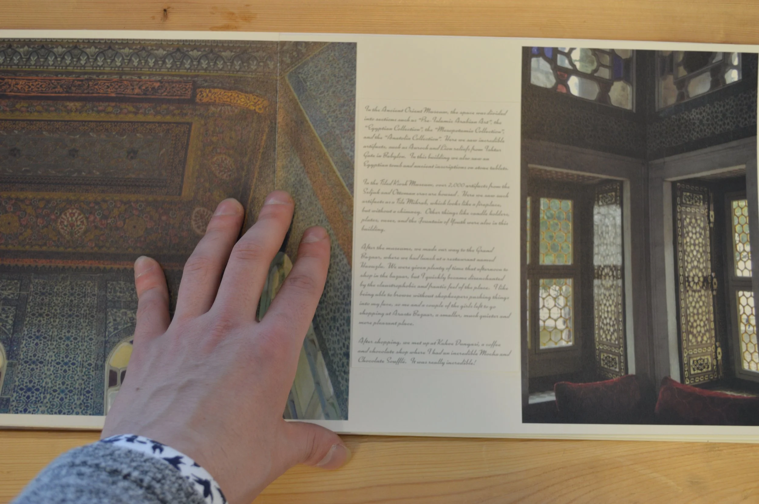 A person with a patterned sleeve is holding a booklet open, showing photographs of intricately decorated rugs and architectural interiors, along with a paragraph of text on the page.