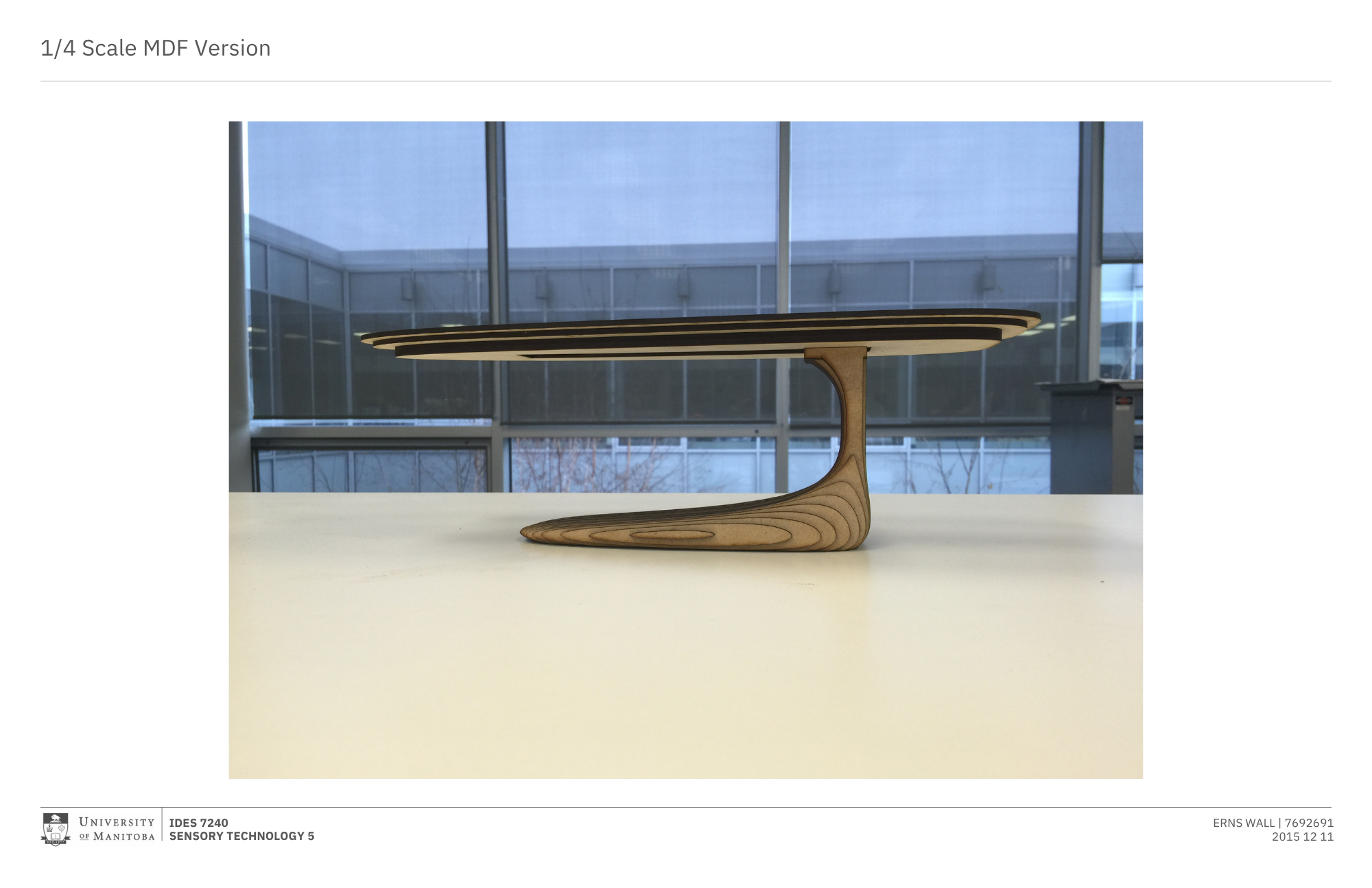 A wooden model of a modern, minimalist, and curved table placed in front of large  glass windows, with an overcast sky and a building visible outside.
