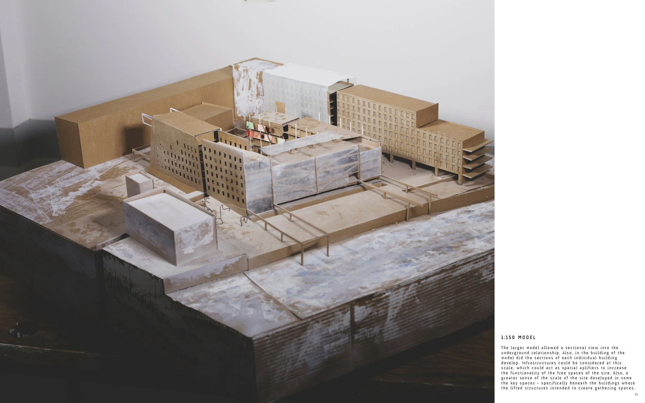 Architectural model of a large building complex with multiple structures, towers, and open spaces, made primarily of cardboard and transparent materials, set on a wooden base.