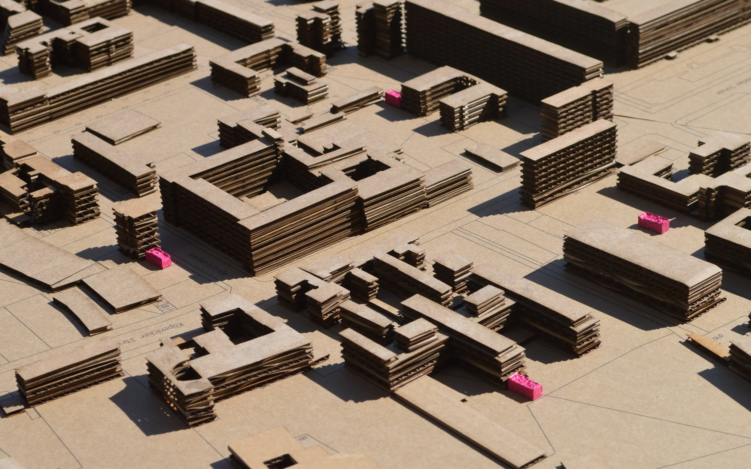 A detailed architectural model composed of stacked pieces of cardboard, representing a cityscape with multiple buildings and structures on a blueprint base.