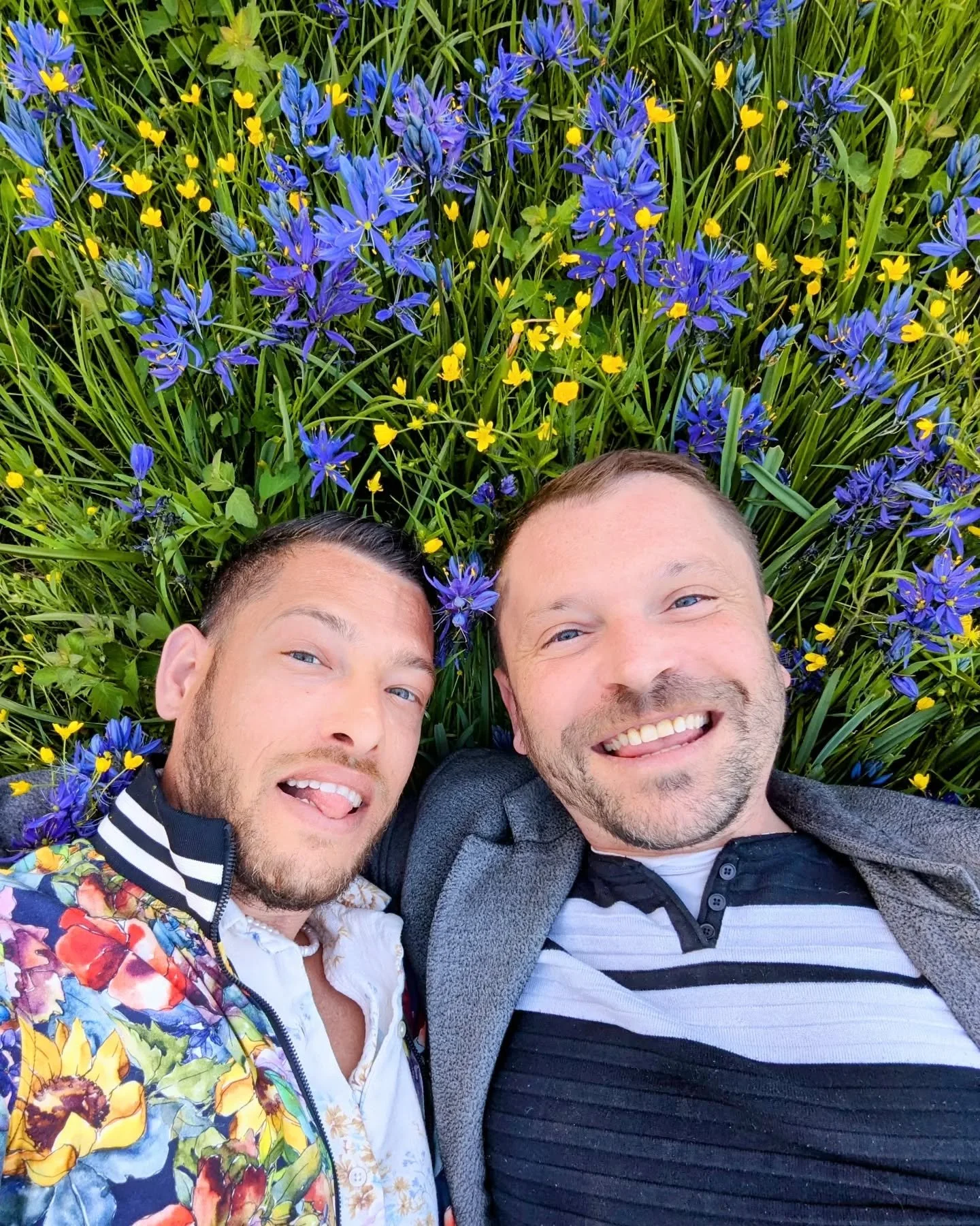 Erns and David lying on the grass surrounded by blue and yellow wildflowers, smiling at the camera.