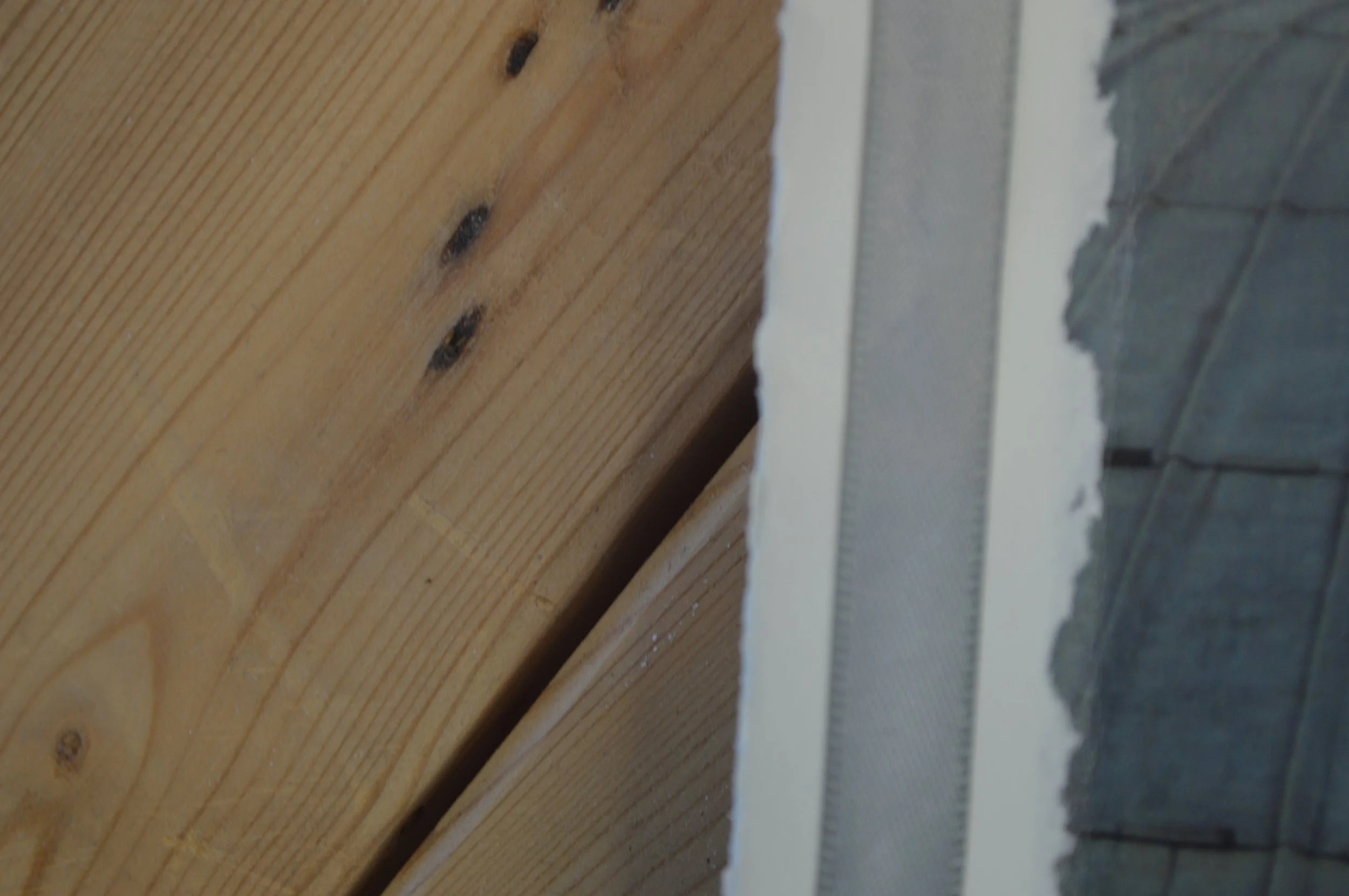 Close-up of a wooden surface with natural grain and dark knots, alongside a section of a brick wall painted gray, separated by a white painted surface.