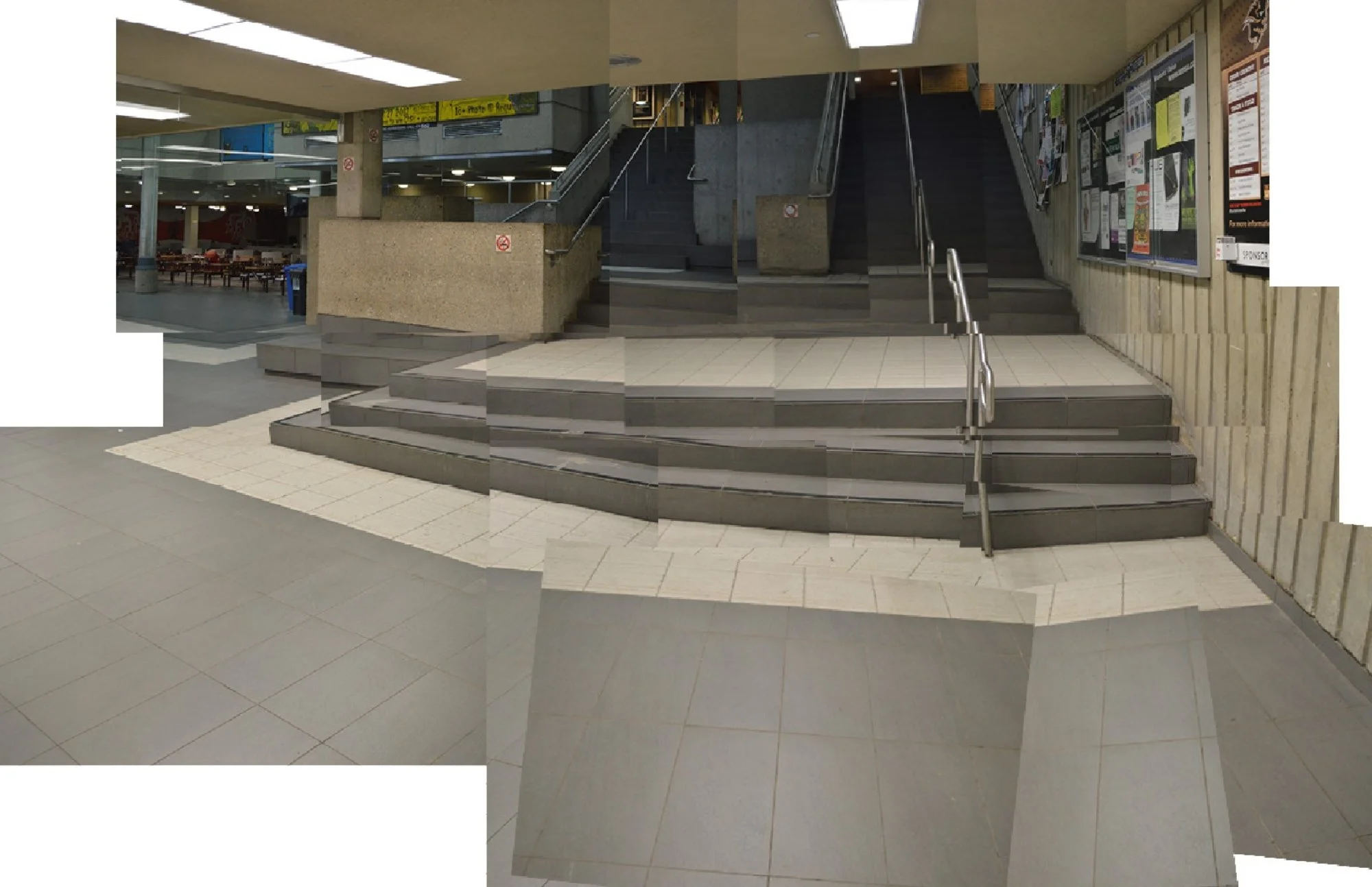 Inside a building with an escalator, stairs, and tiled flooring, featuring a bulletin board with various posters on the wall.