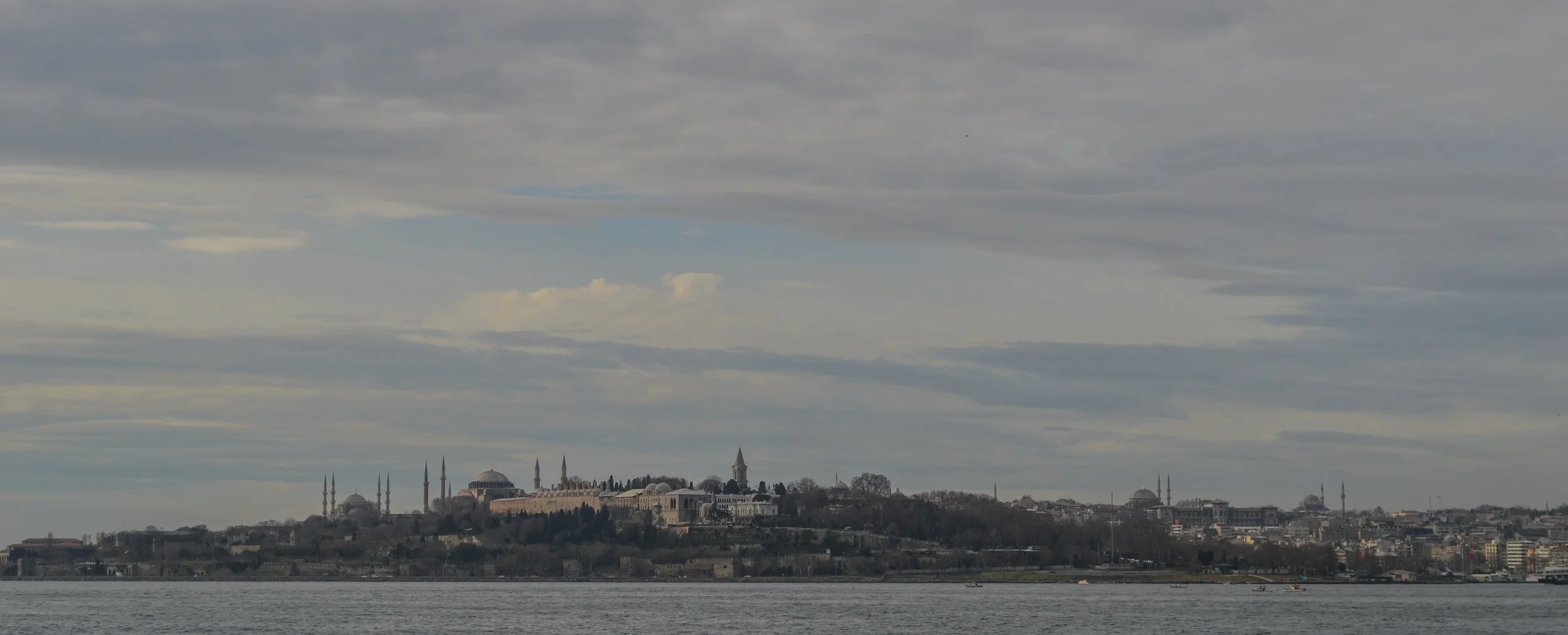 A city skyline across a body of water with a large historic building complex featuring a mosque with multiple minarets and domes, under a cloudy sky.