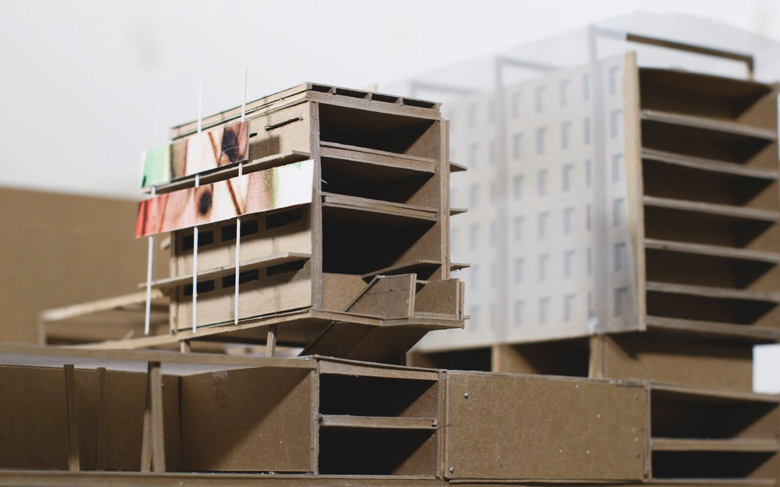 A detailed architectural model of a building structure made from brown cardboard, featuring multiple floors and open spaces, with some colorful printed elements attached to the exterior.