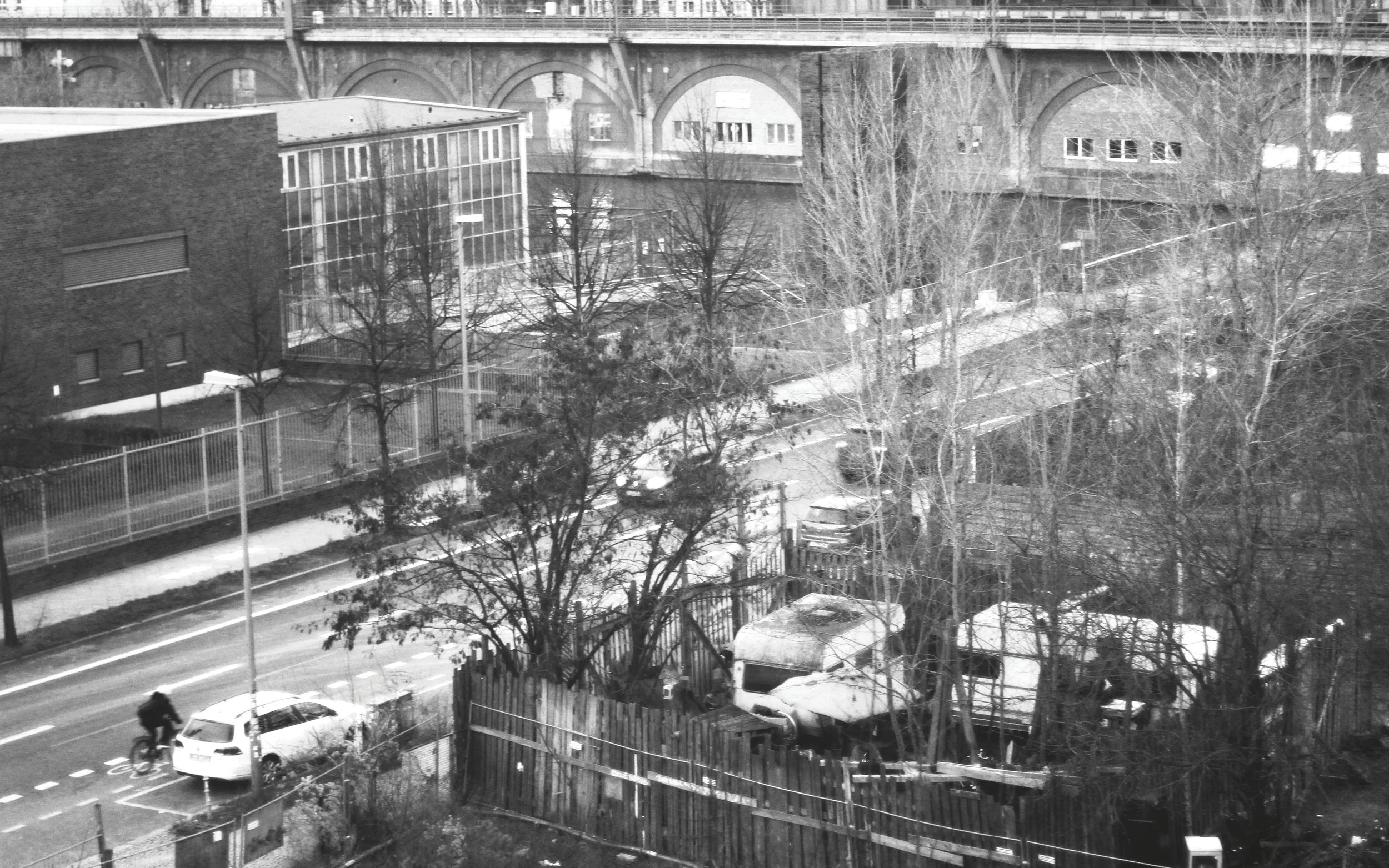 Black and white photo of an urban scene with a street, parked cars, a cyclist, leafless trees, and buildings, with a railway bridge overhead.