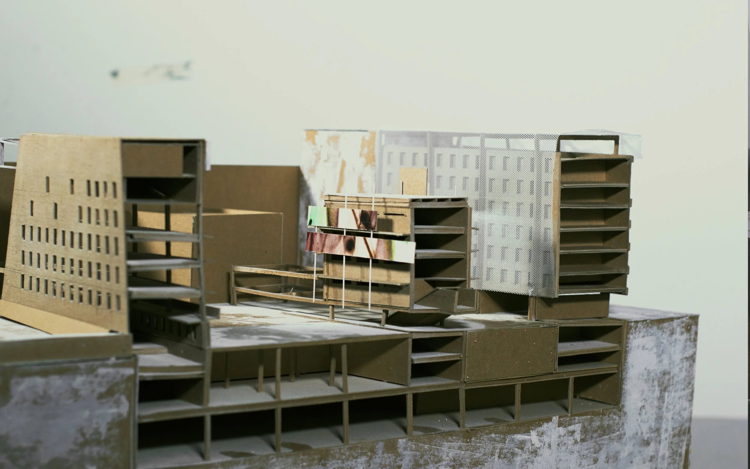 Scale model of a modern architectural building with multiple levels, open windows, and a mix of solid and perforated surfaces, set on a table with a plain wall in the background.