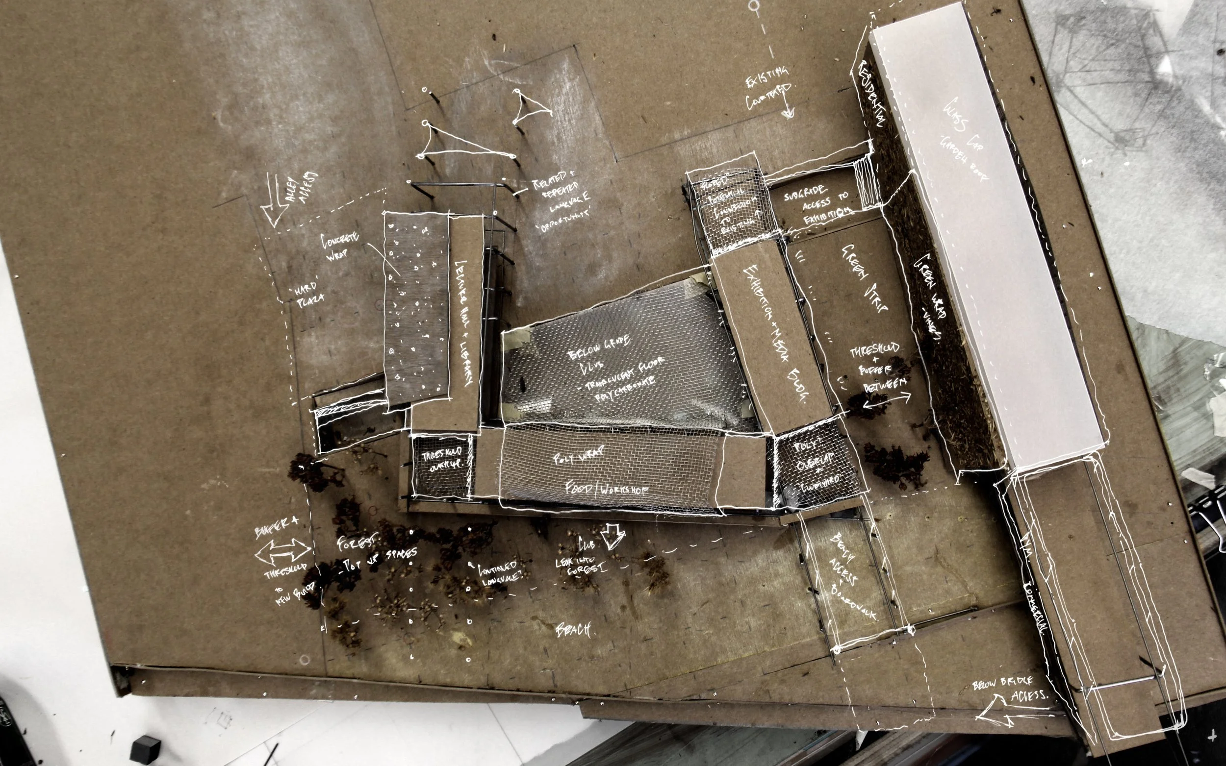 A top-down view of a construction or architectural plan drawn on a surface with white ink, illustrating various sections of a building including a foyer, workshop, and outdoor areas, with annotations and measurements.