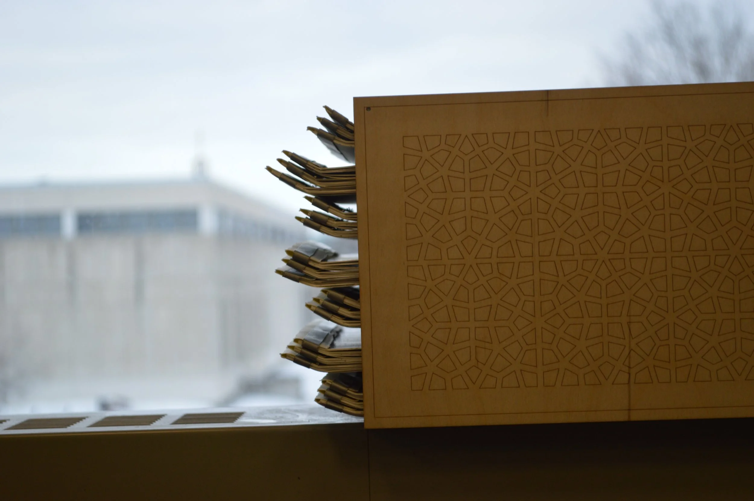 A wooden desk with a geometric cutout pattern, stacked with paper documents, positioned in front of a window with a blurry cityscape outside on a cloudy day.