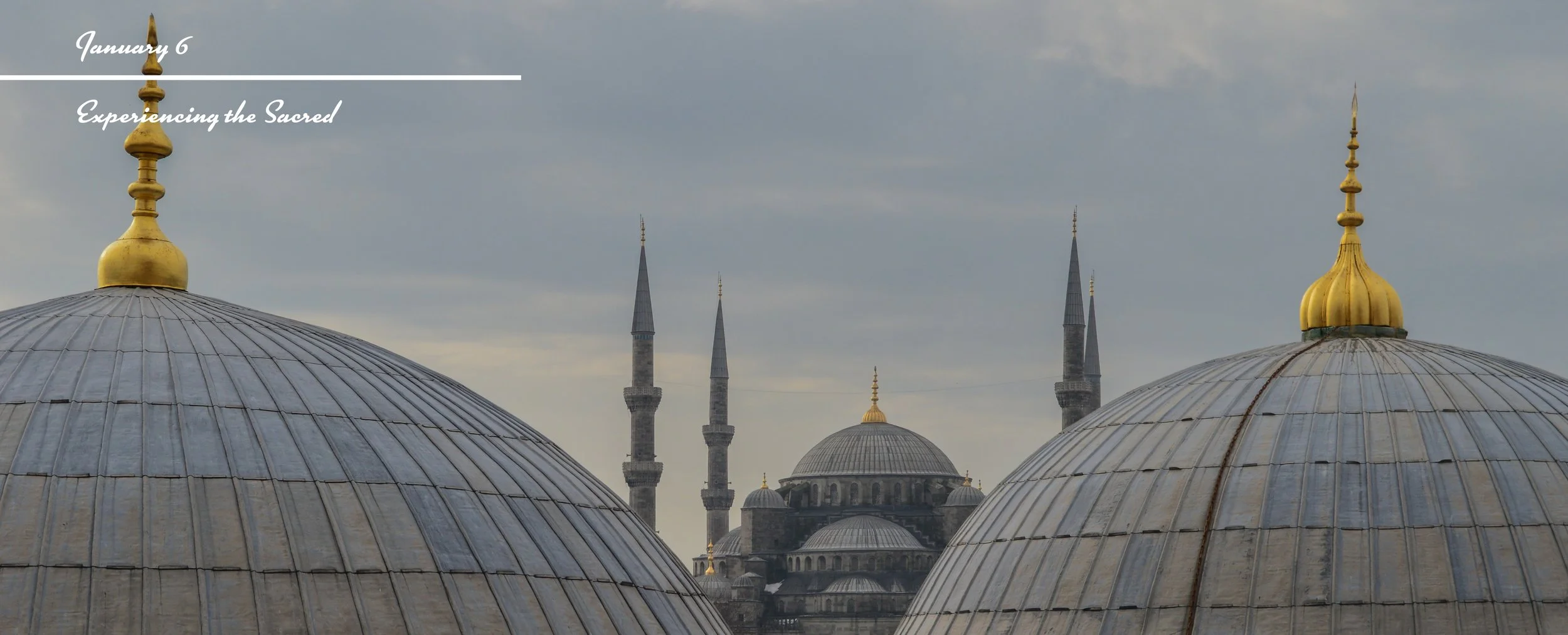 View of domed roofs with gold finials and a mosque in the background, with tall minarets and a cloudy sky.