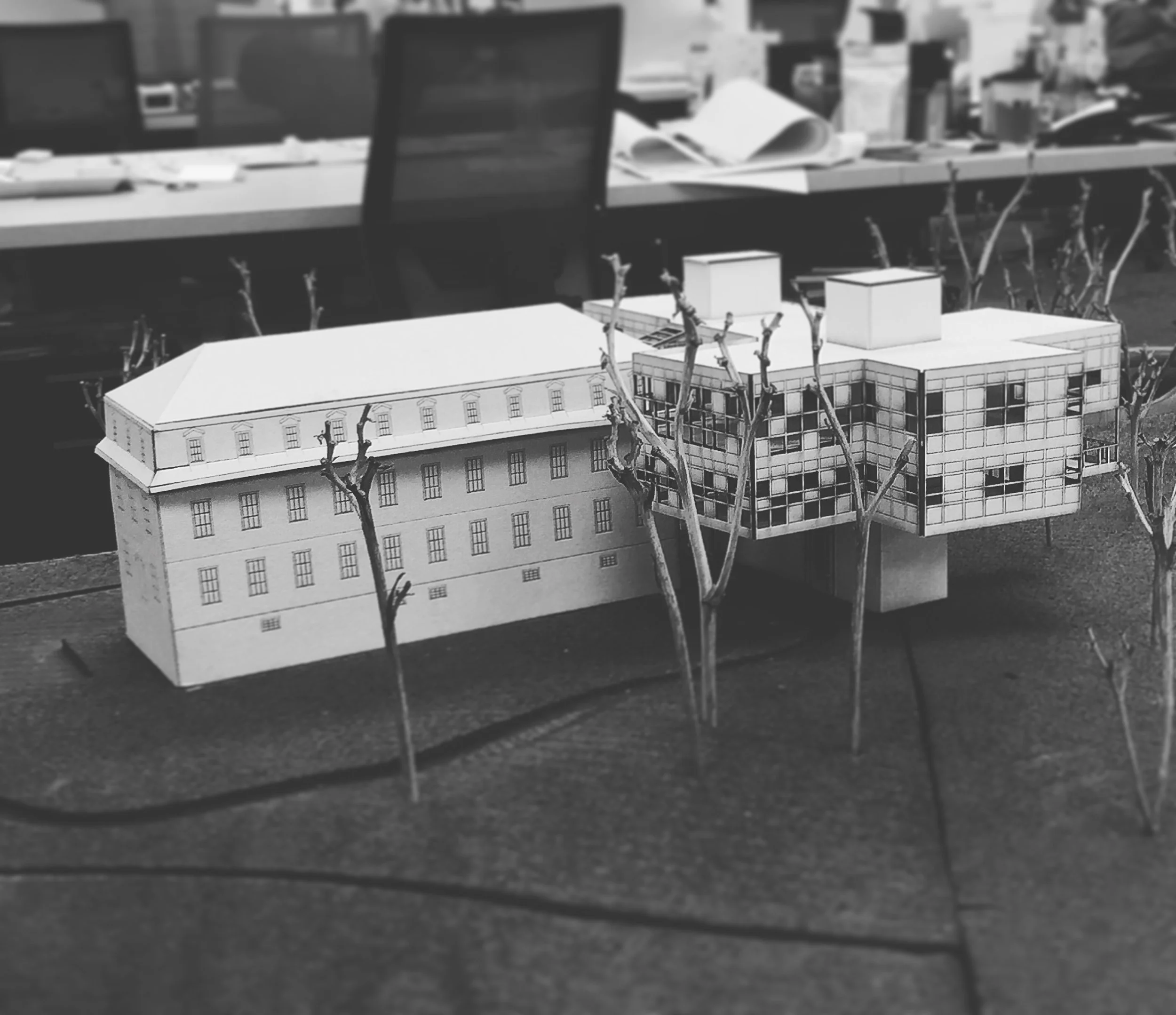 Black and white image of an architectural model of a multi-story building with windows, leaning against a dark surface, with small, leafless trees in front of it.
