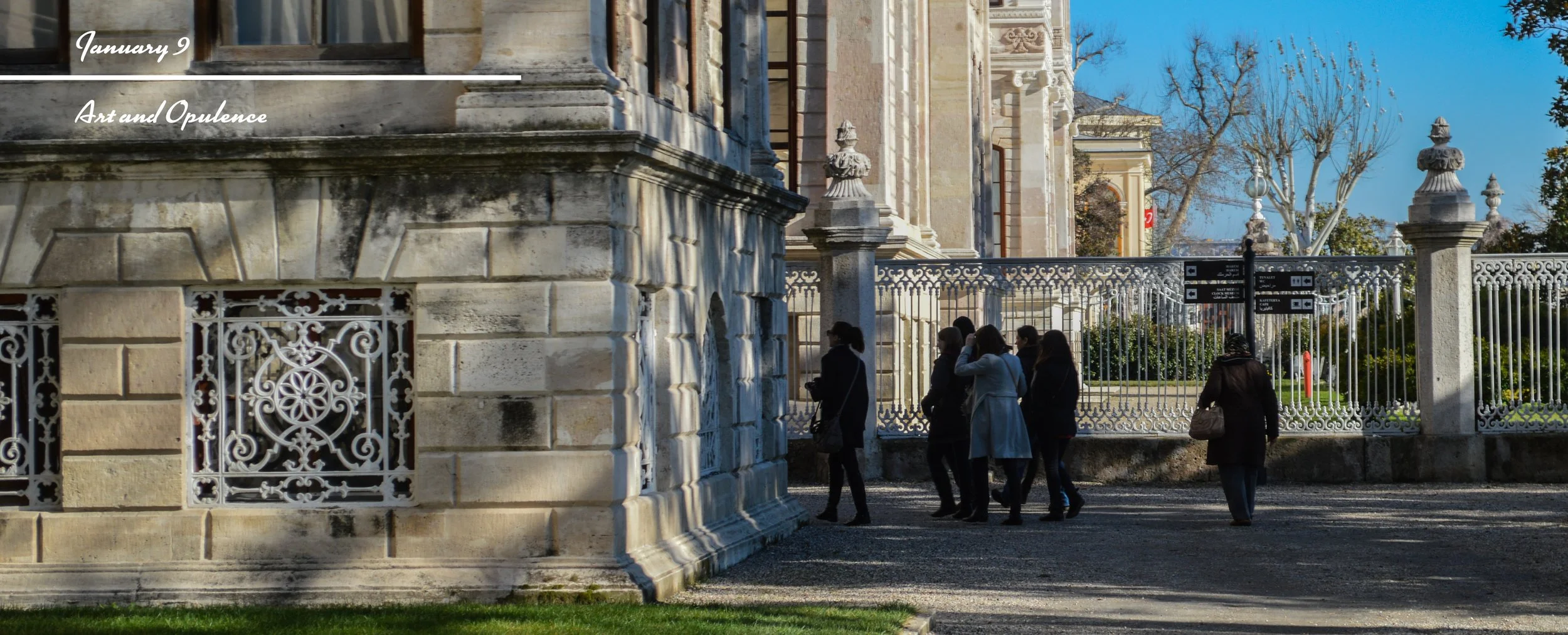Group of people walking past a historic building with ornate wrought-iron fence and stone architecture, on a sunny day with blue sky and leafless trees in the background.