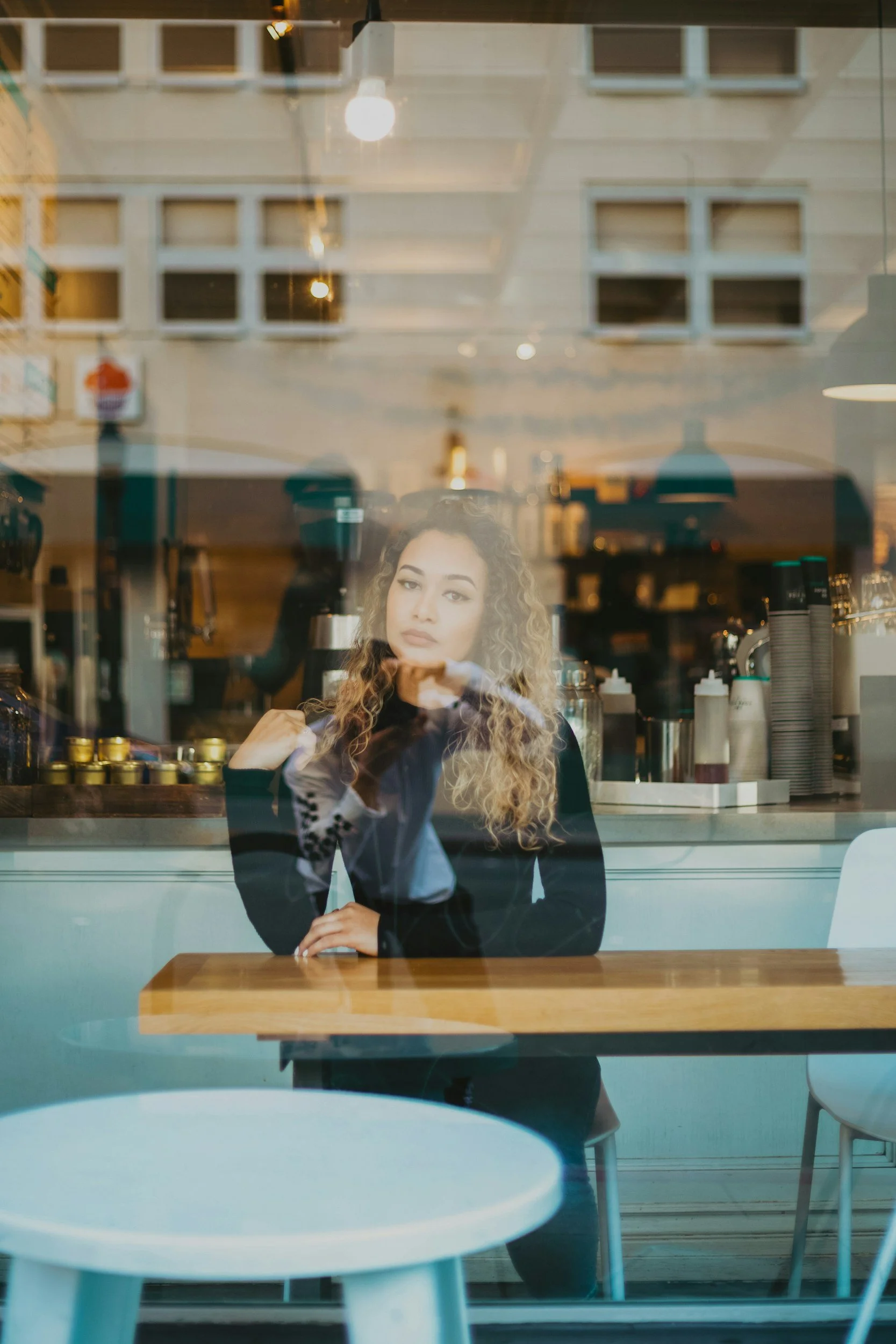 Woman sitting in a quiet coffee shop looking thoughtful while others socialize around her, representing the internal experience of social anxiety.