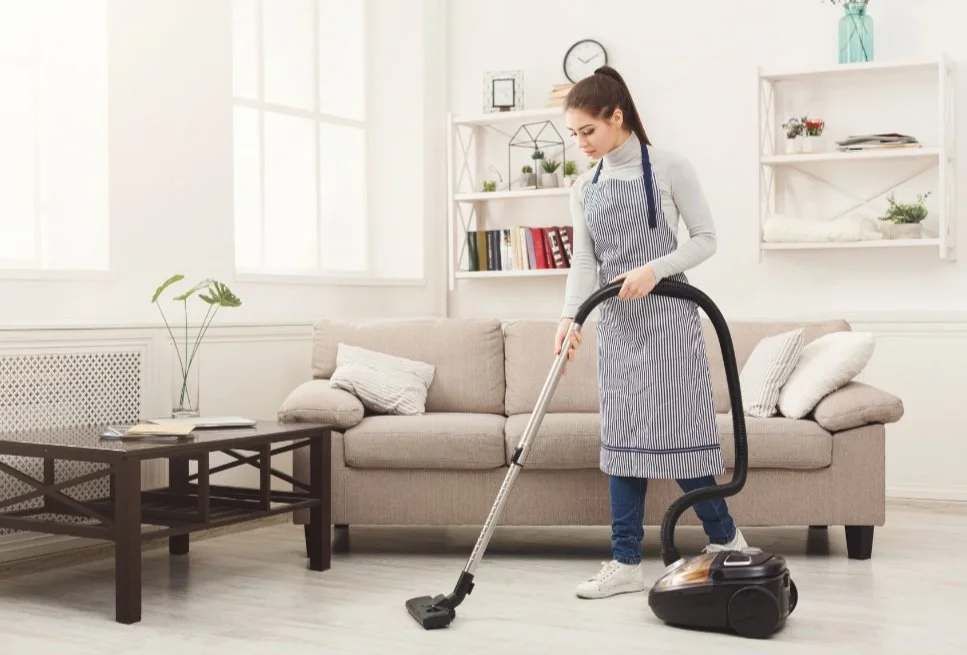 Woman vacuuming in a living room with a beige sofa, white walls, and shelves with books and decorations.
